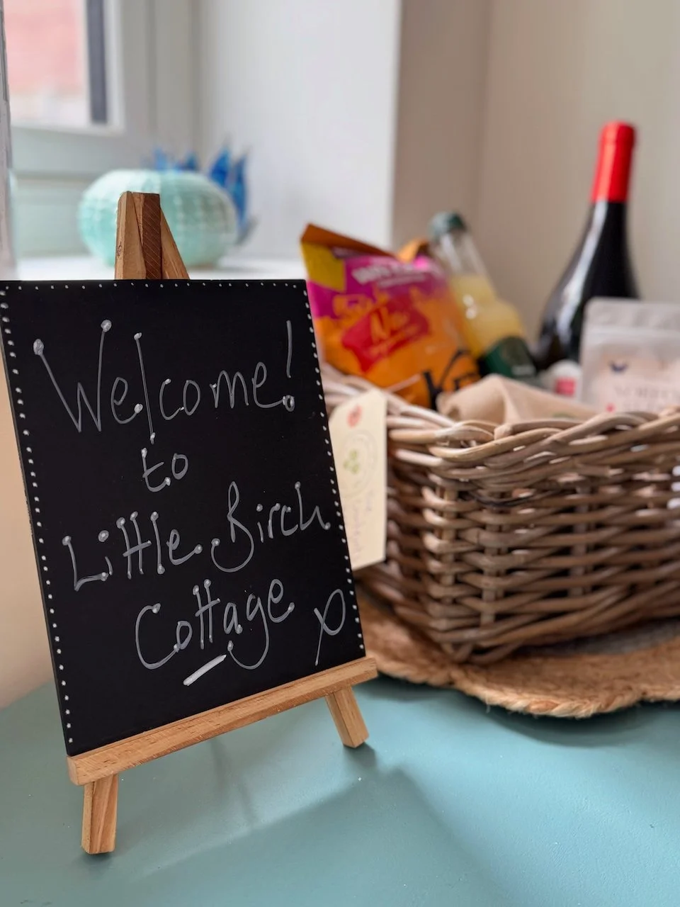 A small chalkboard sign on a table that reads 'Welcome to Little Birch Cottage'—part of a welcoming setup at a cottage, with a wicker basket filled with food and drinks in the background.