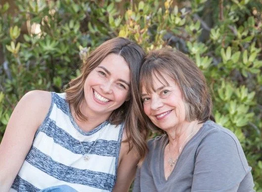 Two women smiling outdoors in front of green bushes.