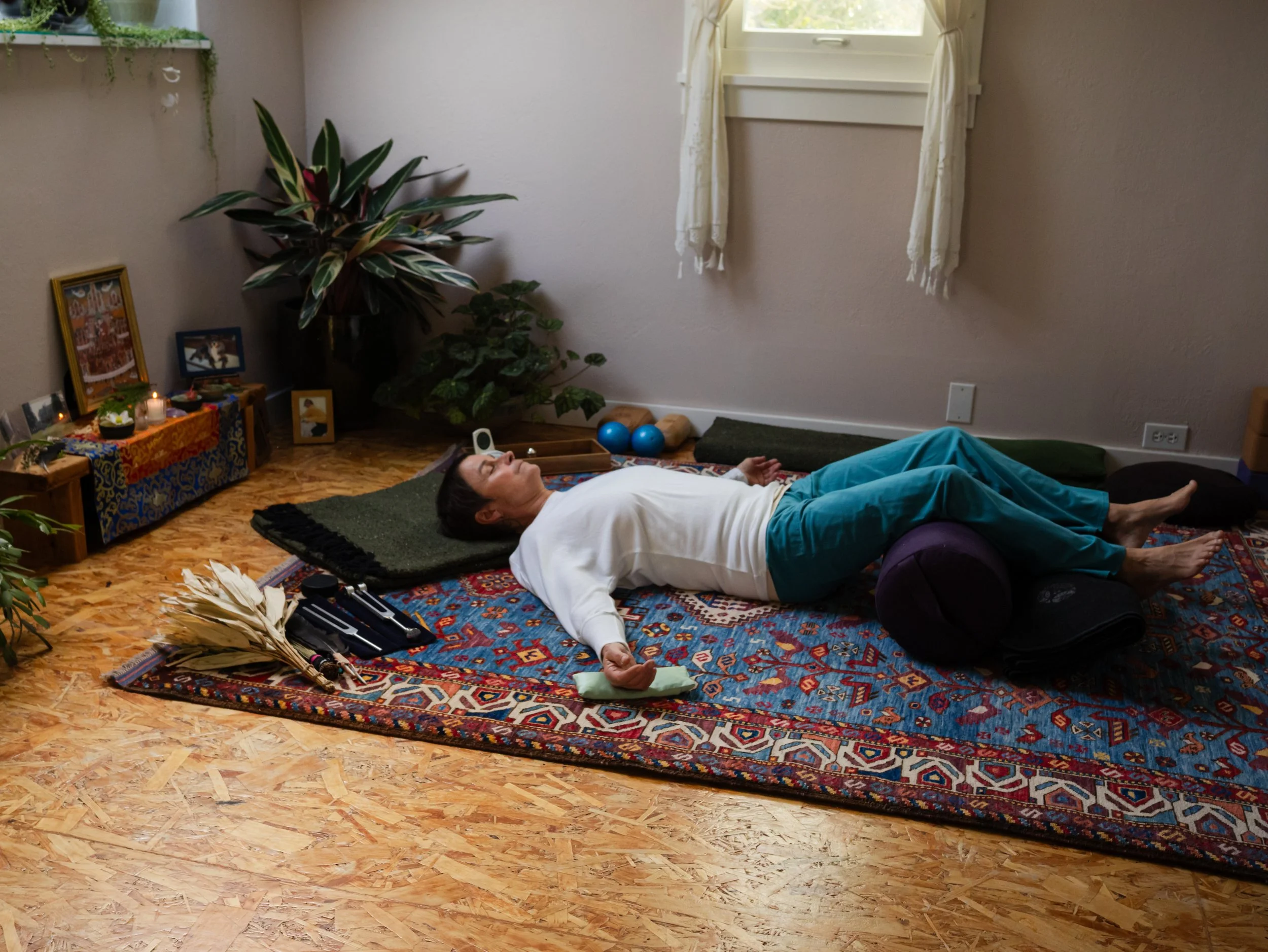 A person lying on their back on a colorful patterned rug in a cozy room with a large houseplant, framed photographs, candles, and spiritual items on a small table. The person appears to be meditating or resting with eyes closed, wearing blue pants and a white shirt.