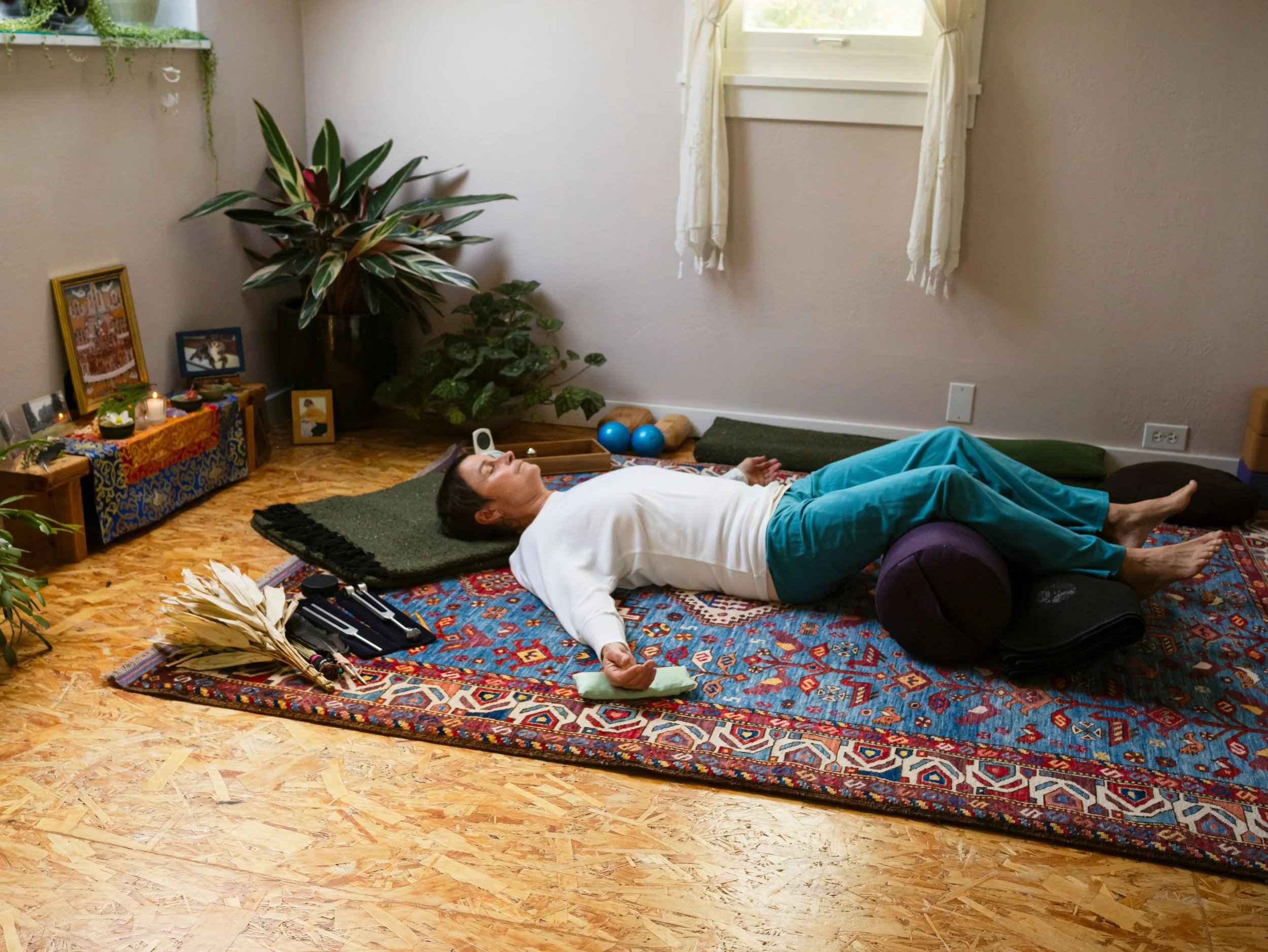 A person lying on a patterned rug on the floor with their eyes closed, appears to be practicing relaxation or meditation. They are surrounded by yoga props like a foam roller, blue exercise balls, and a meditation cushion. A window with curtains lets in natural light, and a shelf with plants and framed pictures decorates the wall.