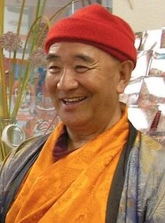 A smiling man wearing a red hat, orange robe, and a dark jacket, standing indoors near colorful plants and shelves.
