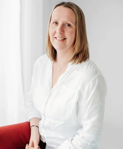 A woman with shoulder-length light brown hair, wearing a white blouse, sitting and smiling in front of a light-colored background.