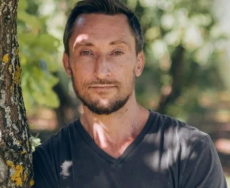 Man with dark hair and beard, wearing a dark shirt, standing outdoors near a tree with sunlight and green leaves in the background.