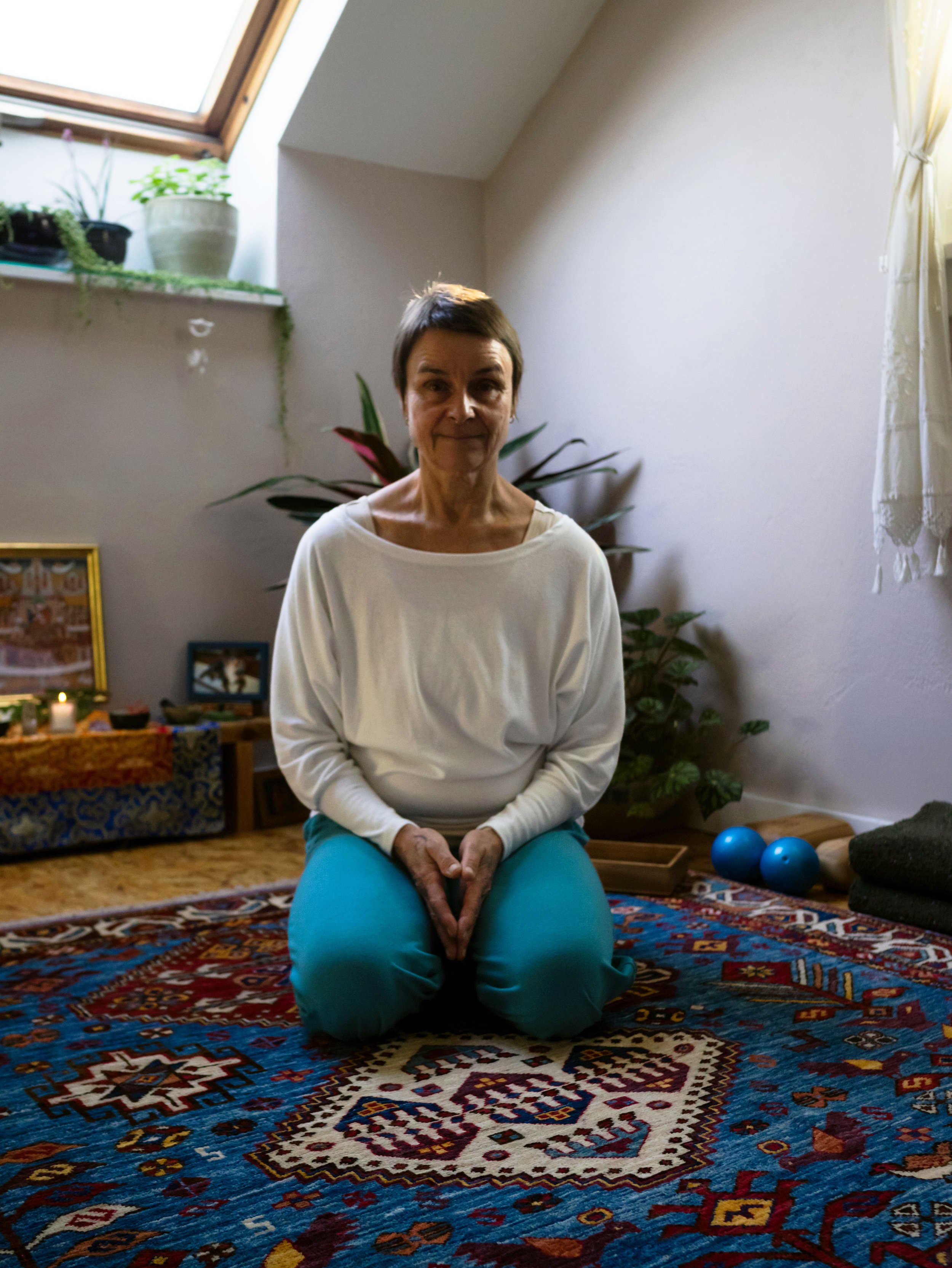 An older woman with short brown hair in a white long-sleeve shirt and teal pants sitting on her knees on a colorful Persian rug in a cozy room with a sloped ceiling and a window, surrounded by plants, framed art, and exercise equipment.