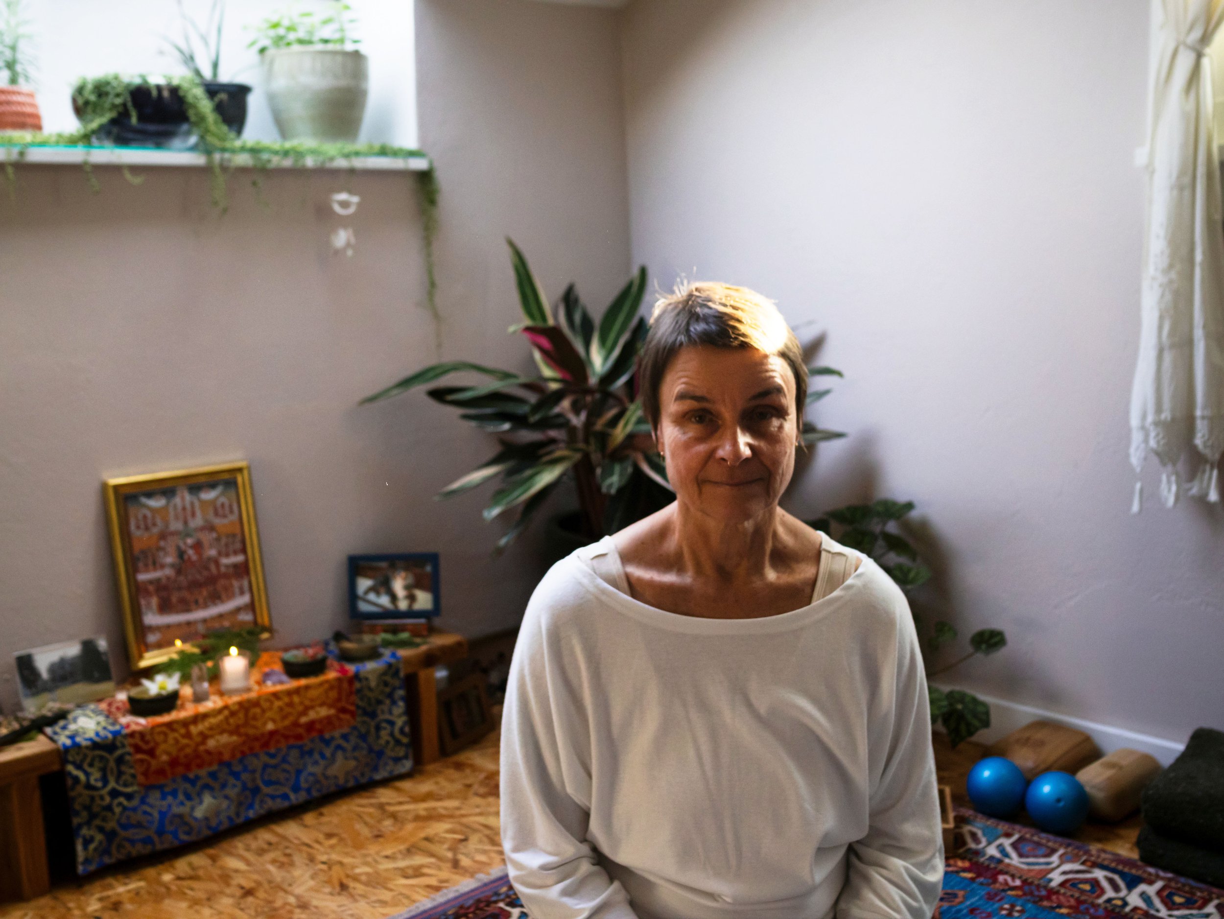 A woman with short hair wearing a white top, standing indoors in front of a plant, with a table filled with colorful items and candles, and framed artwork on the wall behind her.
