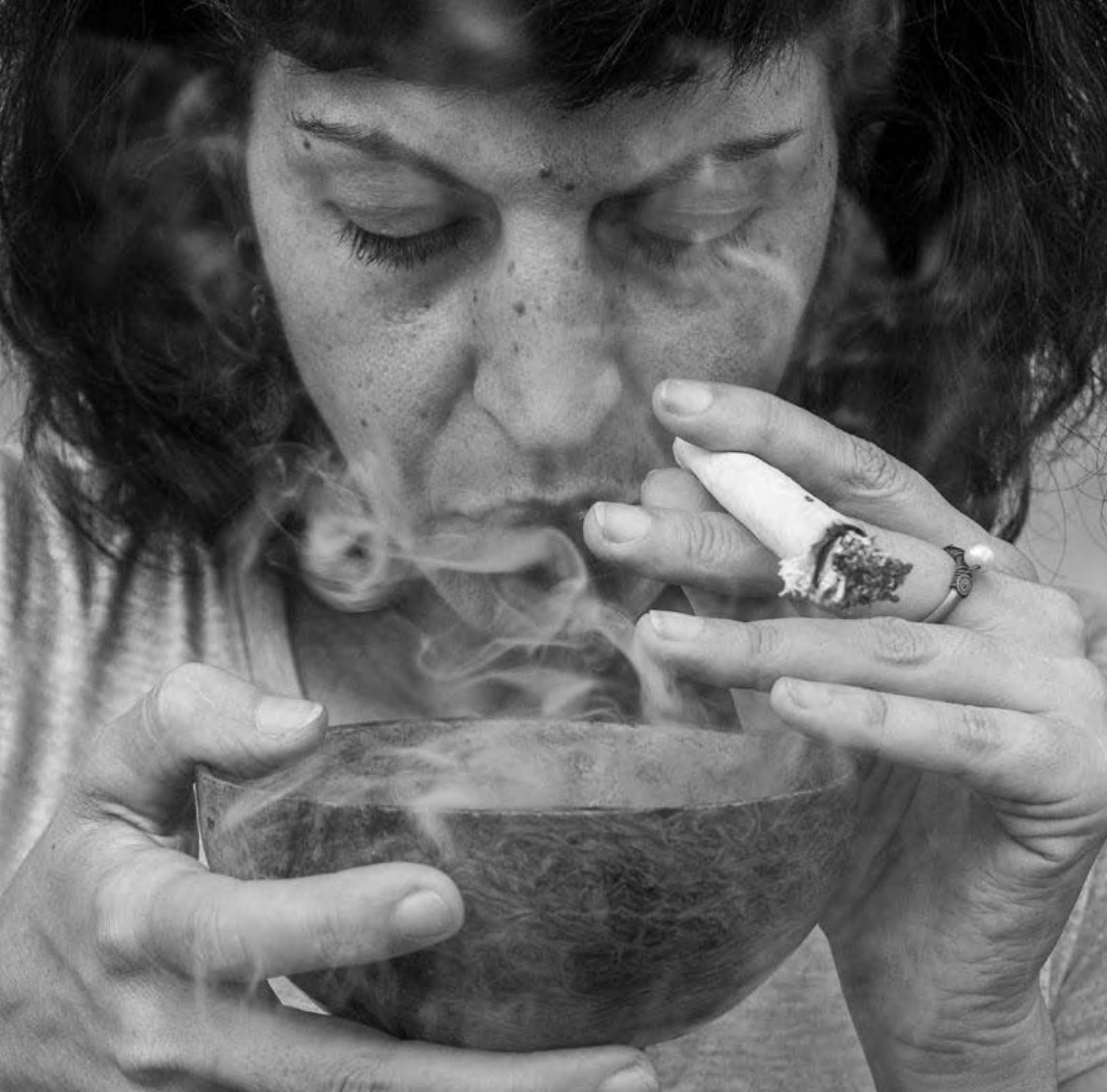 A black and white close-up of a woman with curly hair, eyes closed, holding a steaming bowl close to her face and smoking a cigarette.