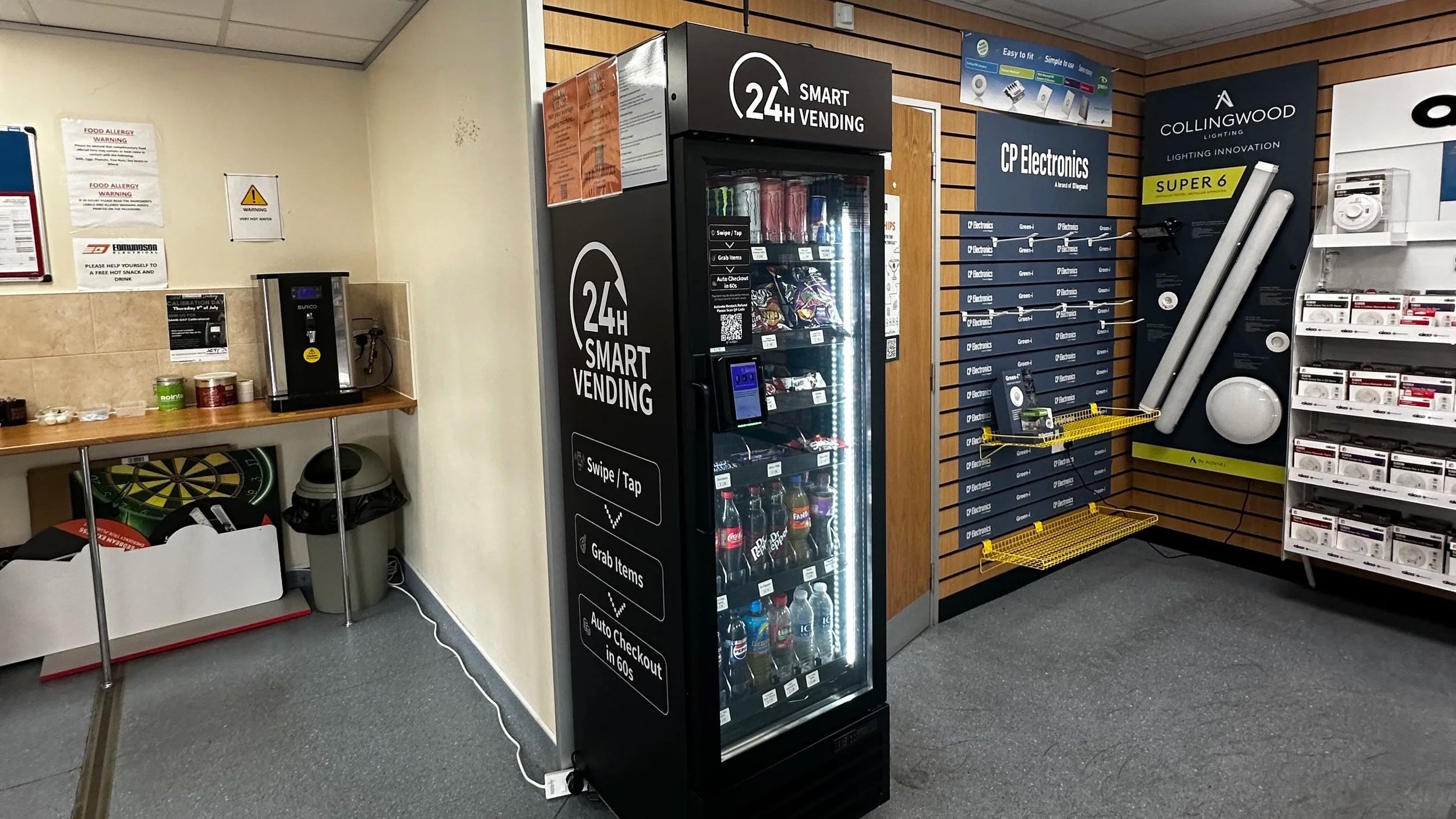 A vending machine labeled '24h Smart Vending' stocked with drinks, a small table with a cardboard box, bottles, and snack items, and shelves with electronic accessories in a retail store.