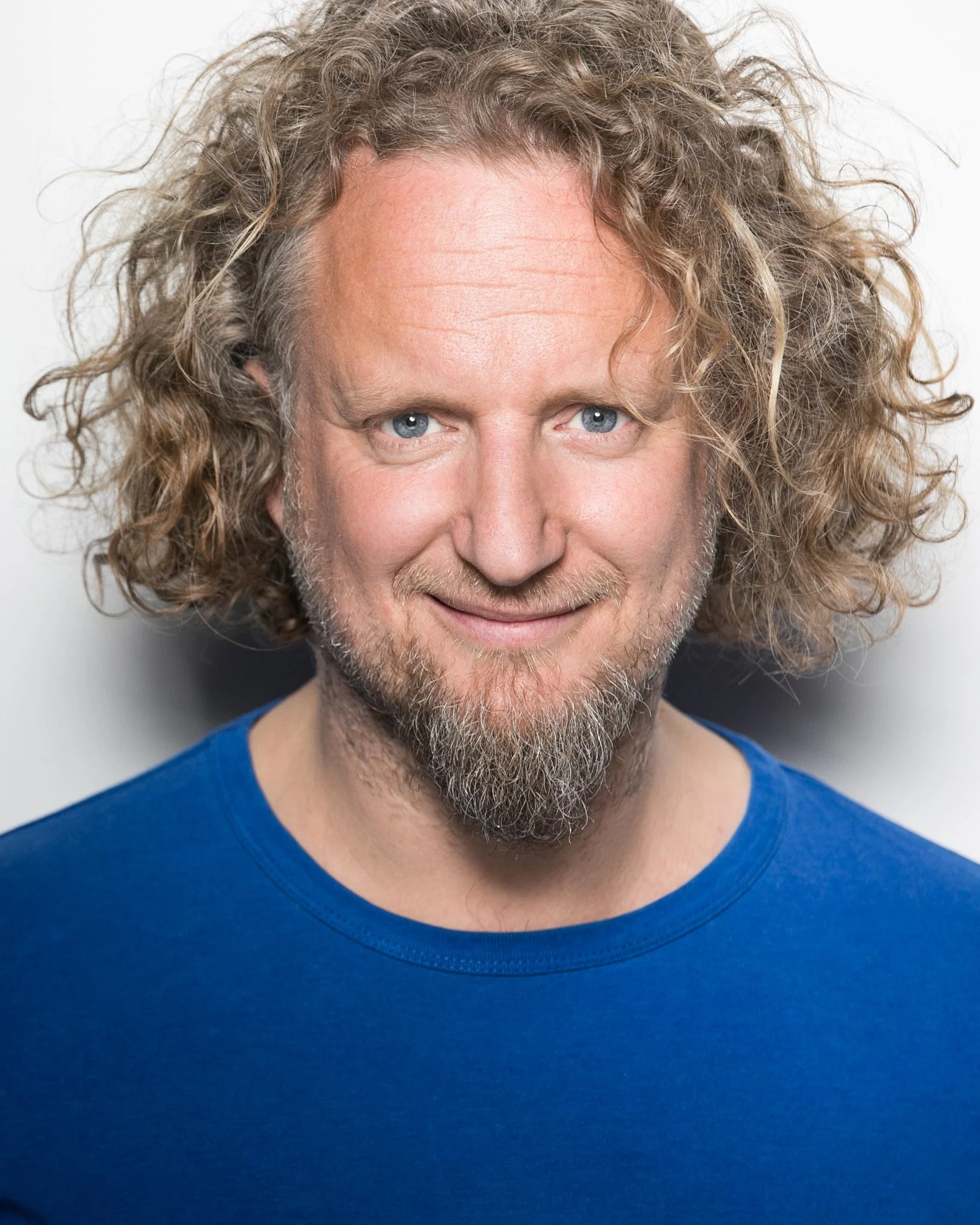 A man with curly blond hair and a beard, wearing a blue shirt, smiling at the camera.