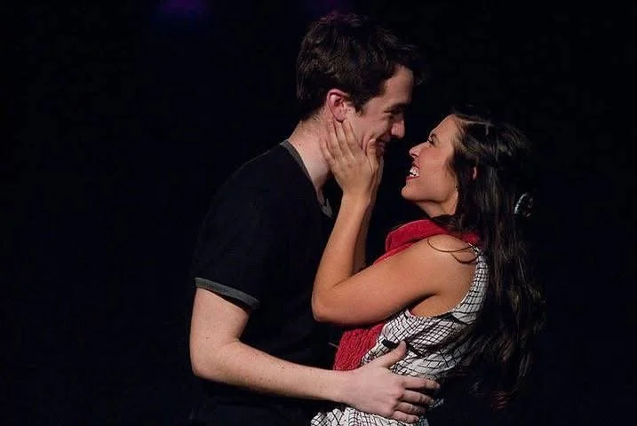 A young man and woman are looking at each other, smiling, with the woman touching the man's face and the man holding her waist.