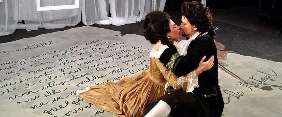 Two actors in period costumes are in an intense embrace, appearing to kiss, on a stage with a large handwritten text background.