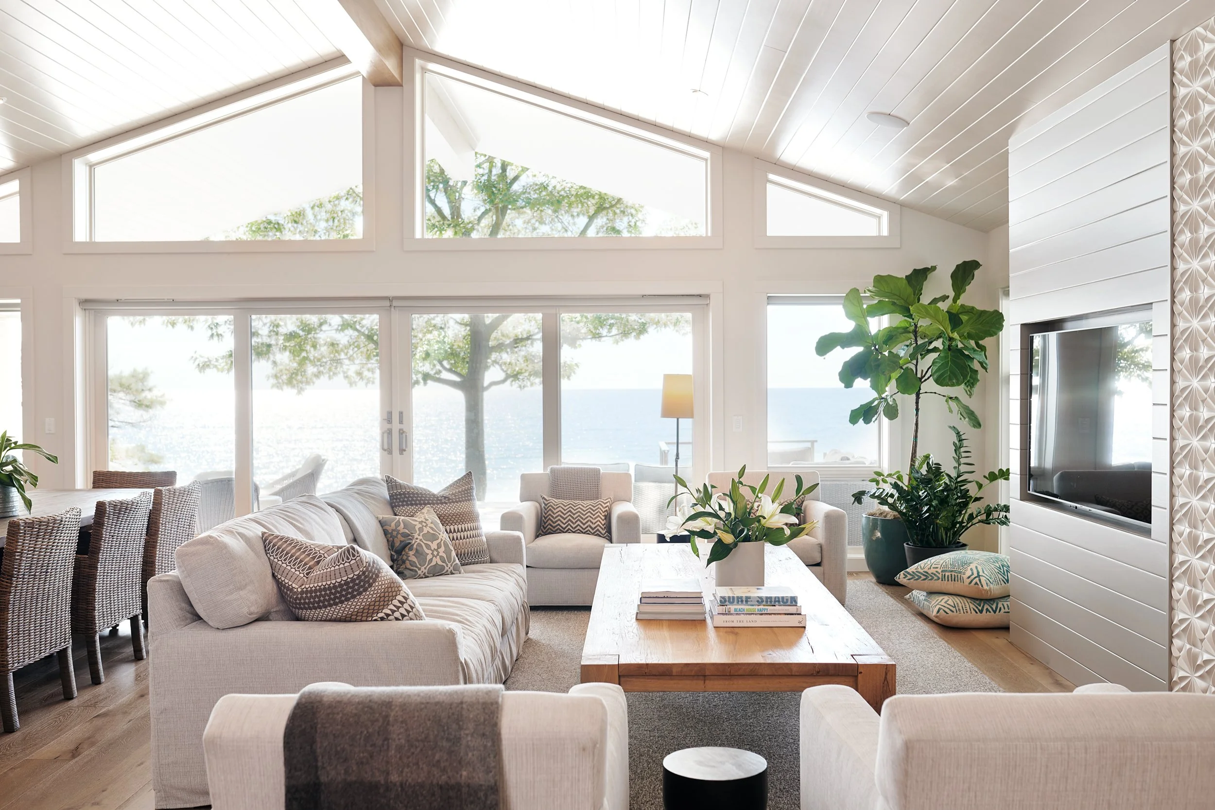 Bright open concept living area with large floor to ceiling windows overlooking Lake Michigan, furnished with sofas, armchairs, a wooden coffee table, and potted plants.