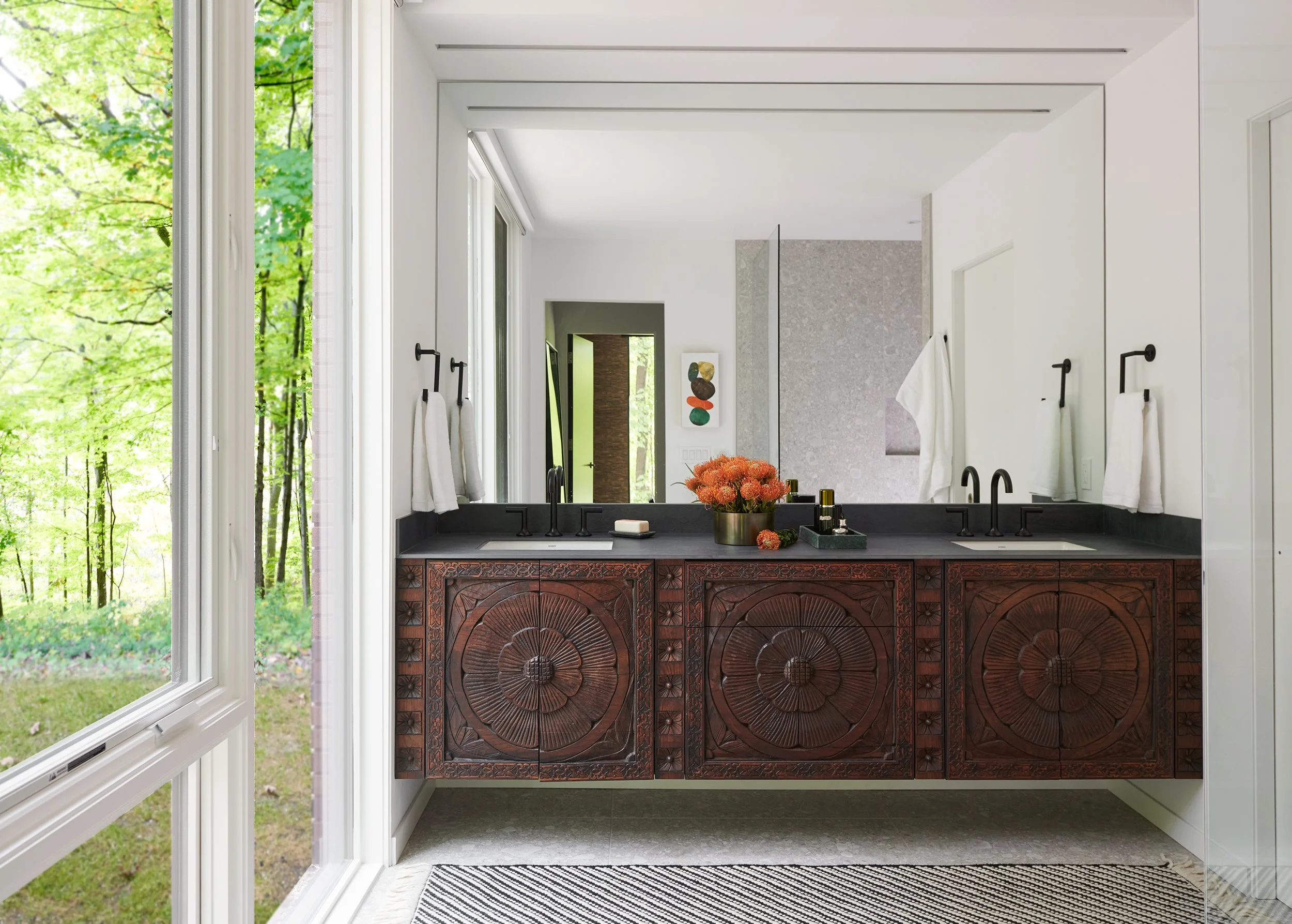 Bathroom with double sinks, dark cabinets with carved floral designs, black faucets, white walls, large mirrored wall, flower arrangement, toiletries, towel hooks with white towels, large window showing a green wooded landscape.