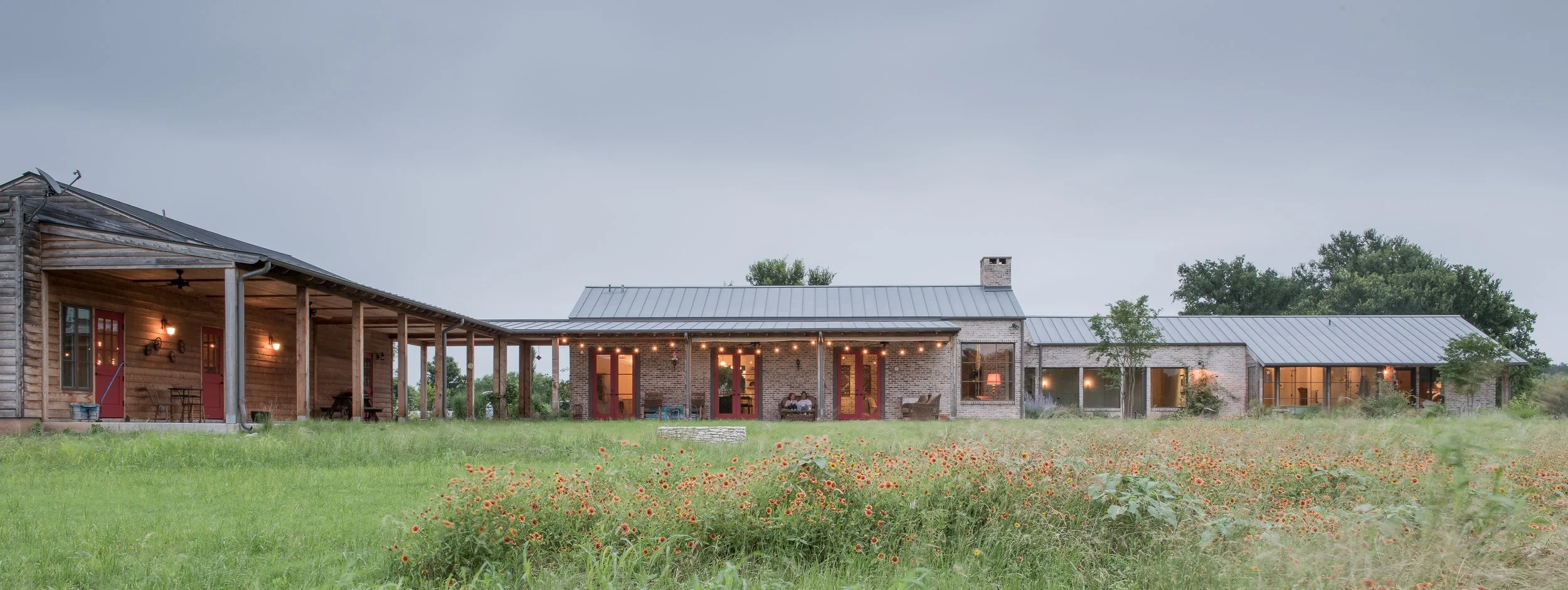 Large rural ranch-style house with metal roof, brick and wood siding, large windows, and a spacious porch area, surrounded by a green field with flowers and trees.