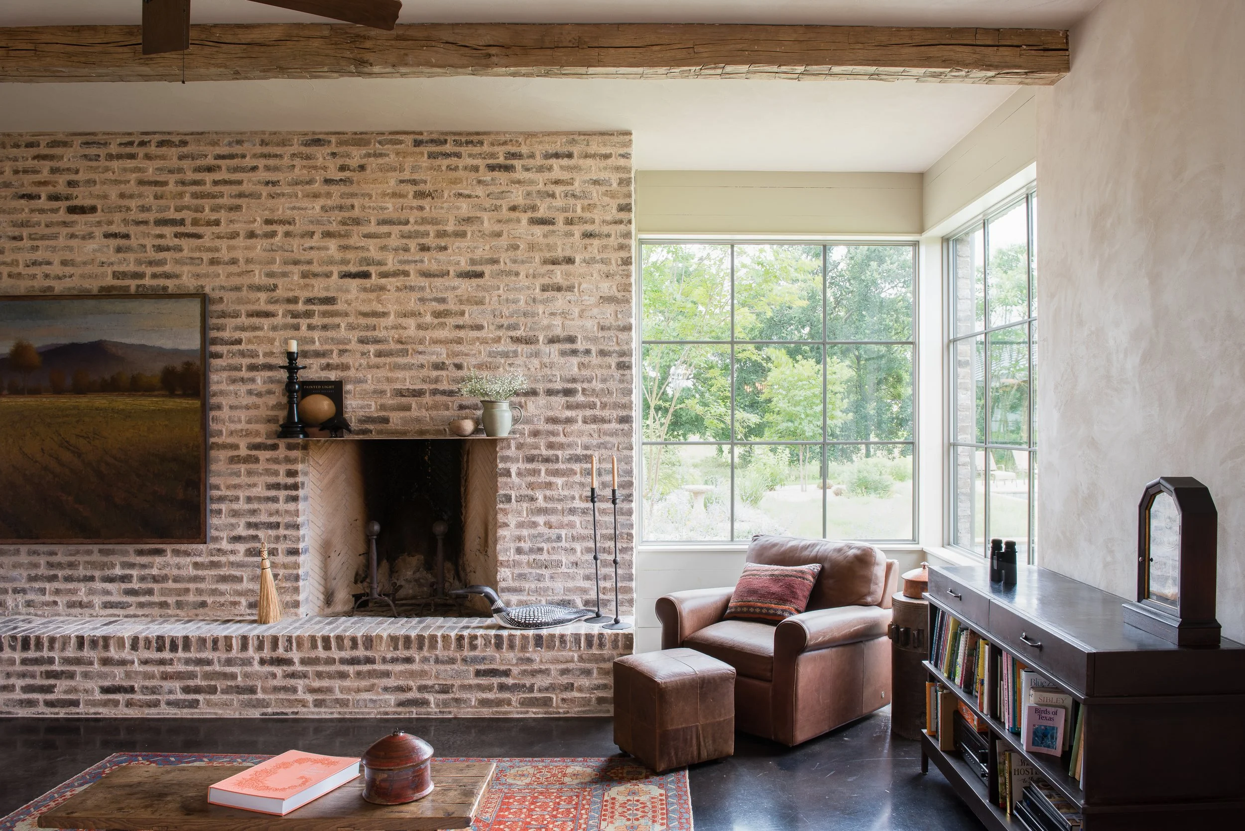 Cozy living room with brick fireplace, leather armchair with pillow, wooden bookshelf, large window with green trees outside, and decorative items on mantle and coffee table.
