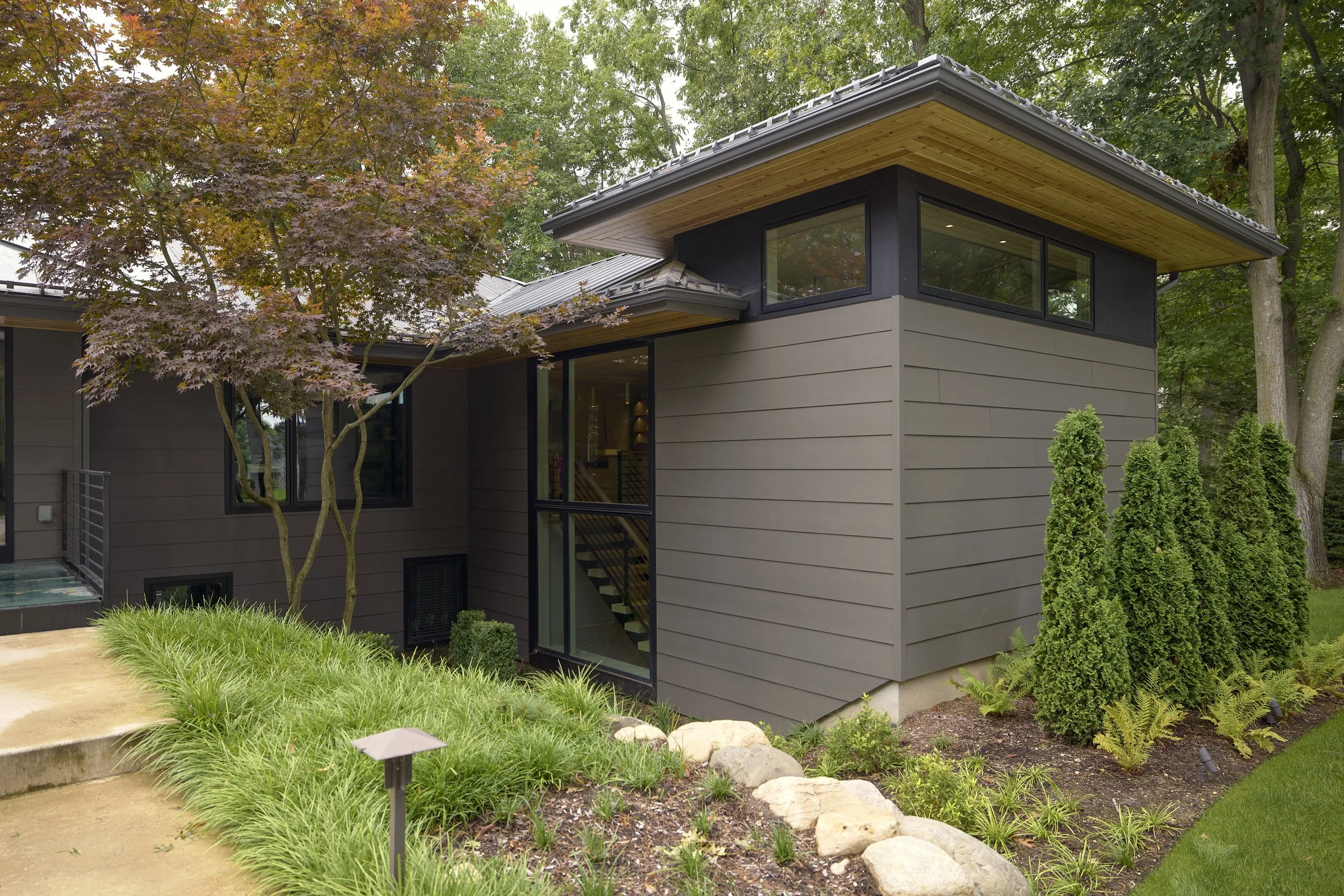 Modern home addition with dark gray exterior, large windows, a wooden overhang, surrounded by lush green trees and landscaped garden with shrubs, grass, and rocks.