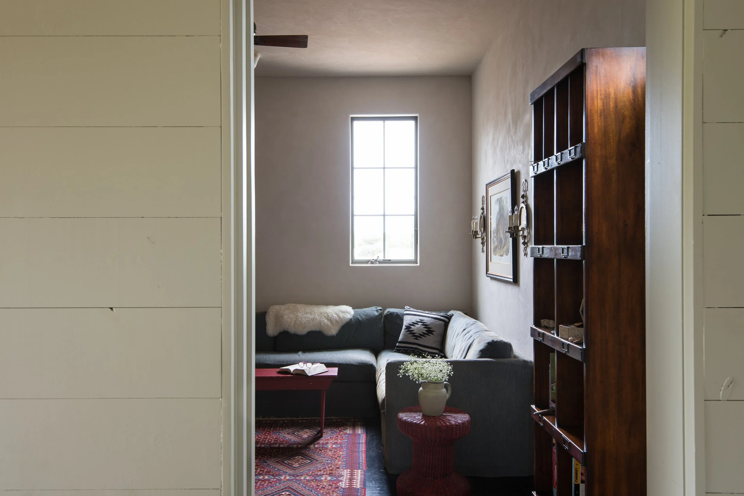 Guest bedroom with a large window, dark gray sectional sofa with patterned pillows, a small red table with an open book, a red woven side table with a potted plant, a wooden bookshelf, wall art, and wall sconces.