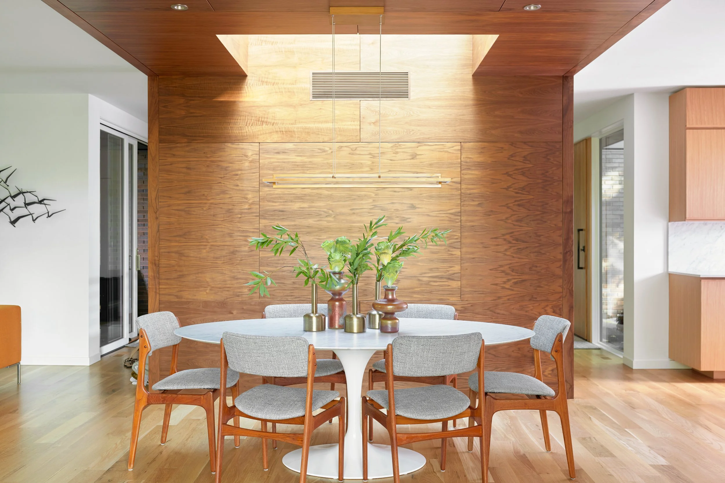 Modern dining room with a round white table topped with greenery in a vase and surrounded by midcentury style chairs. The room features a wood-paneled waterfall wall and skylight. Two pendant lights hang above the table.