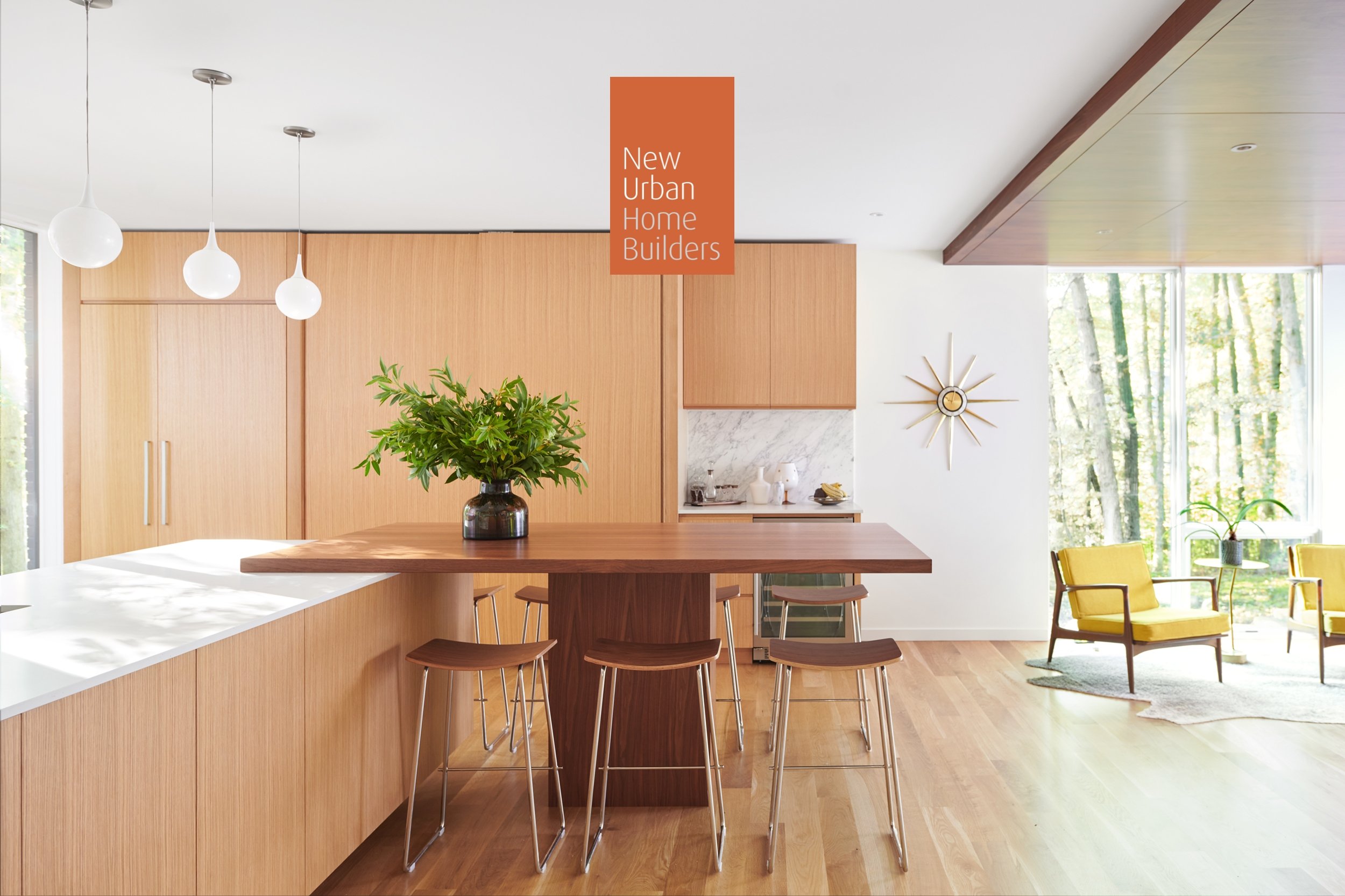 Modern kitchen and living area with custom white oak cabinetry, a central island with a green plant, yellow chairs, and large windows showing a view of trees.