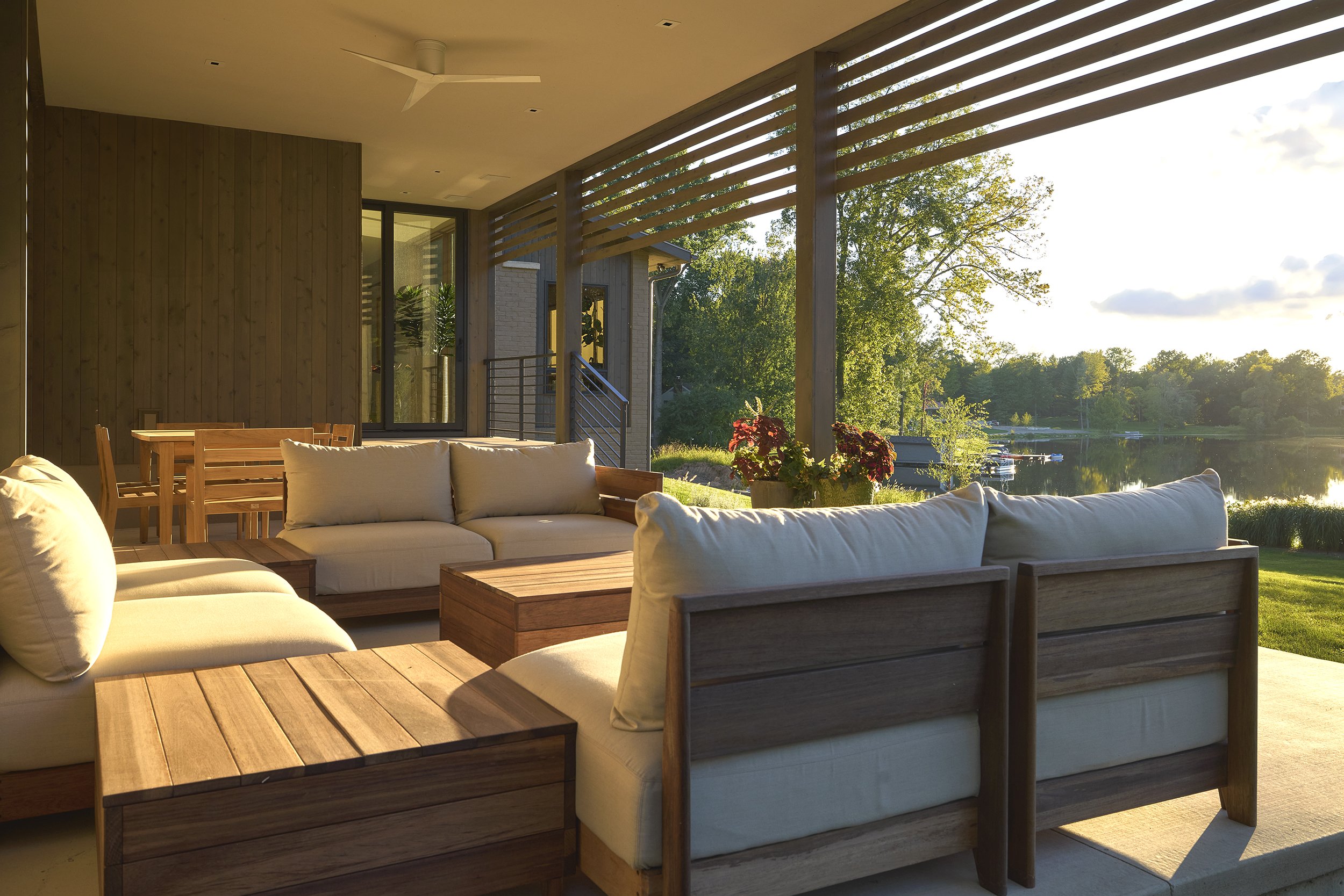 Covered outdoor patio with white cushioned seating, wooden tables, and a view of a lake with trees and dock in the background during sunset.