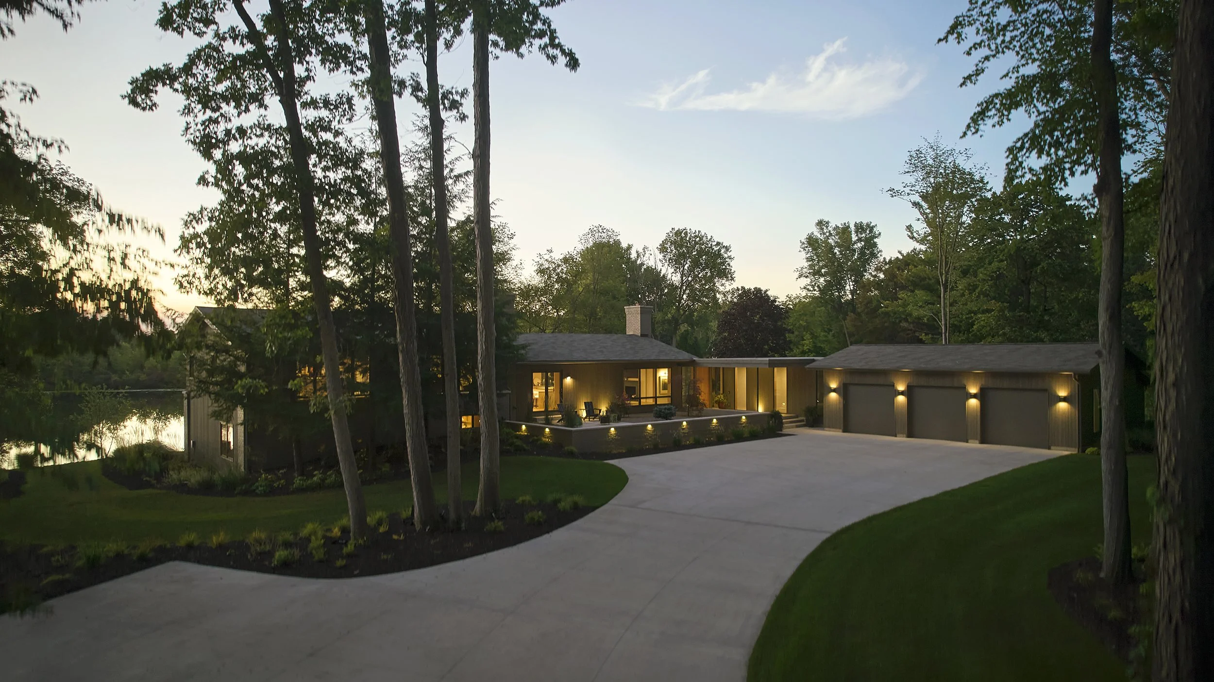 A modern house with a wide driveway, surrounded by trees and greenery, illuminated by exterior lights in the evening.