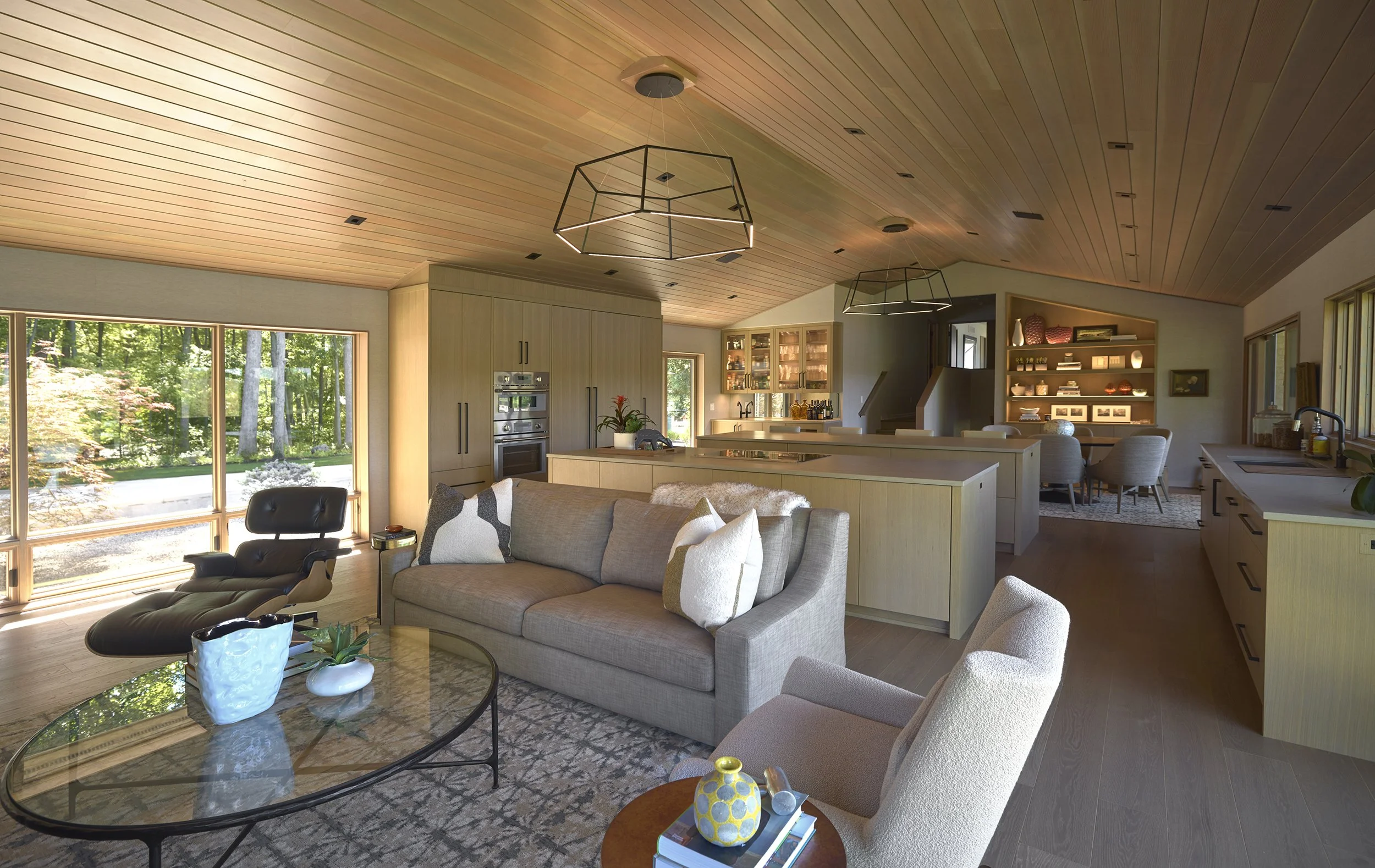 Open concept living area with large windows, light wood flooring, and wood-paneled ceiling contains beige sofa, black lounge chair, glass coffee table. Kitchen with white cabinetry, built-in appliances and dining space in the background.