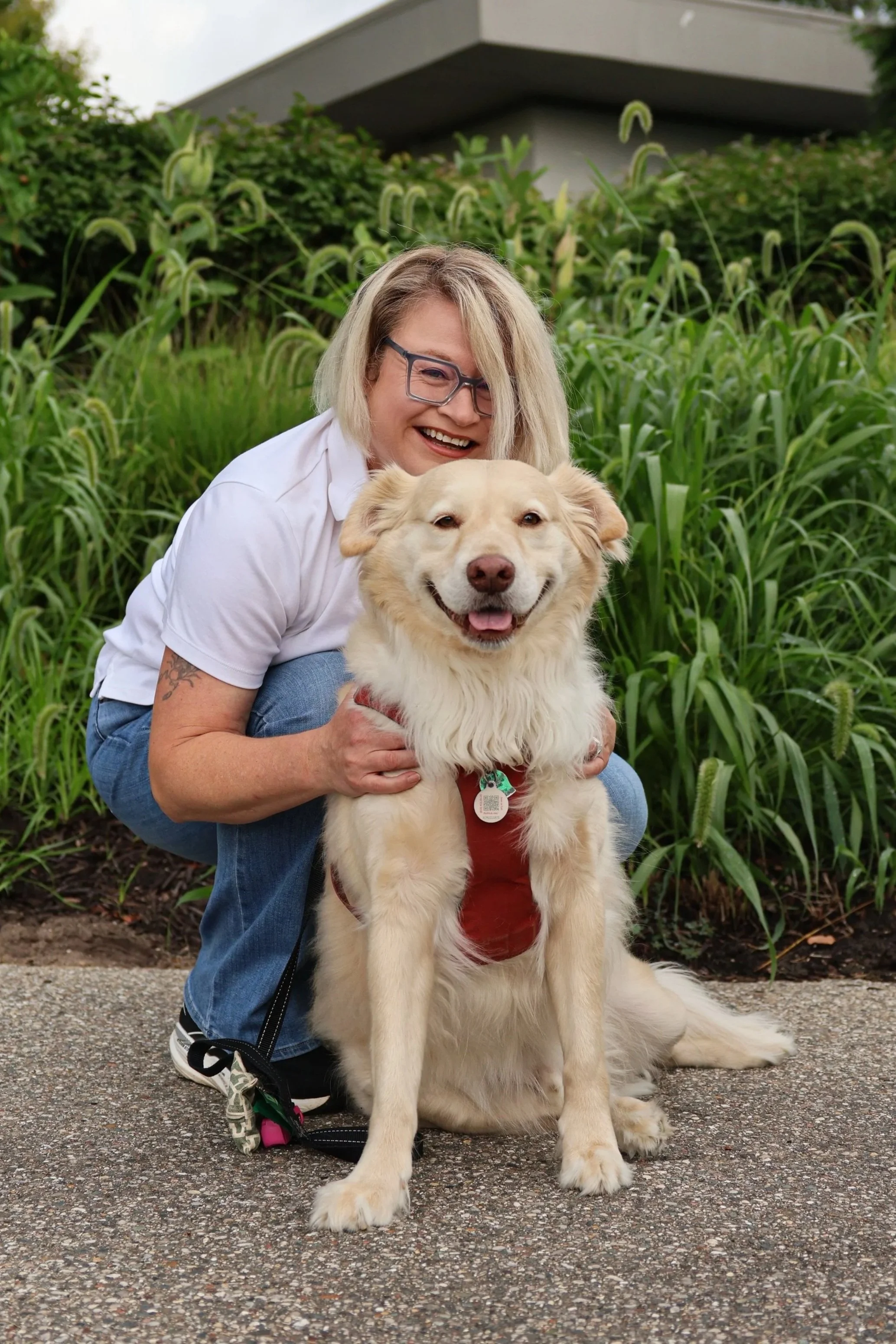 Alison Black crouching with a happy, fluffy Golden Retriever in an outdoor setting with green plants and office building in the background.
