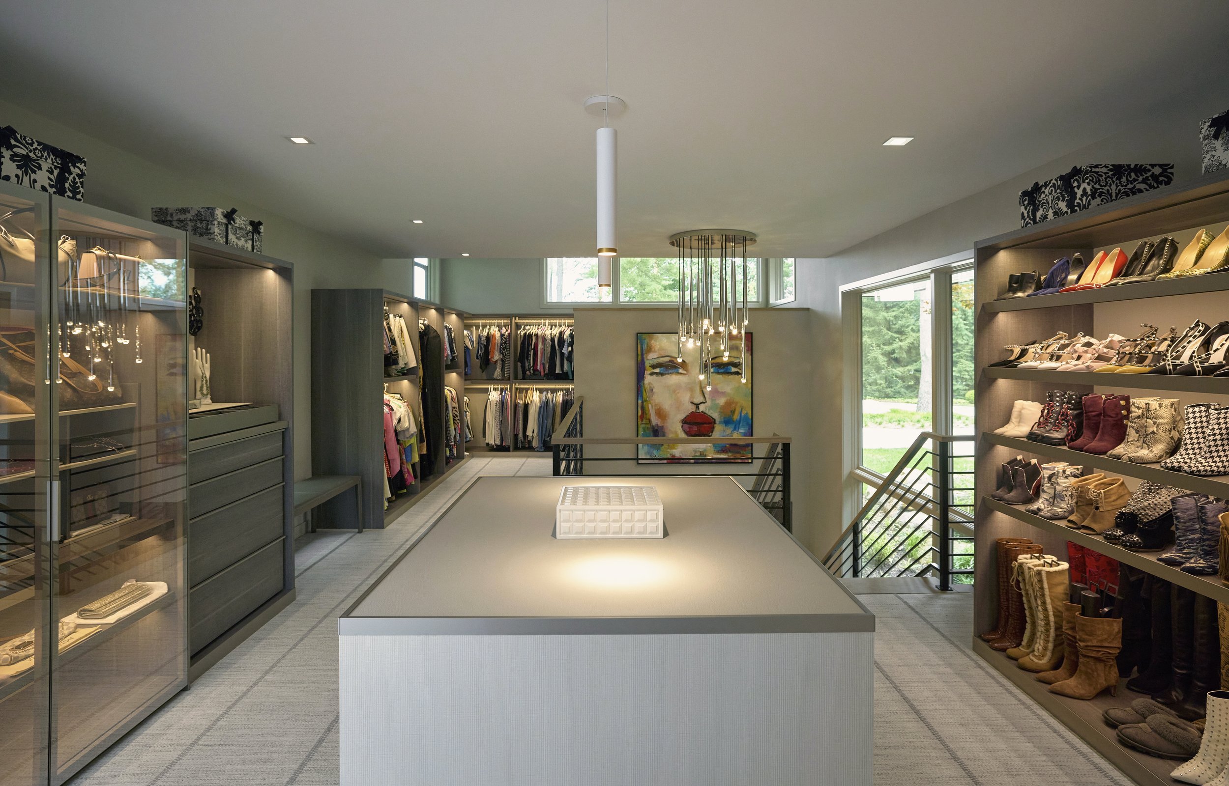 Interior of two story contemporary master closet with custom display shelves, an art piece of a woman's face on the back wall, and large windows showing greenery outside.