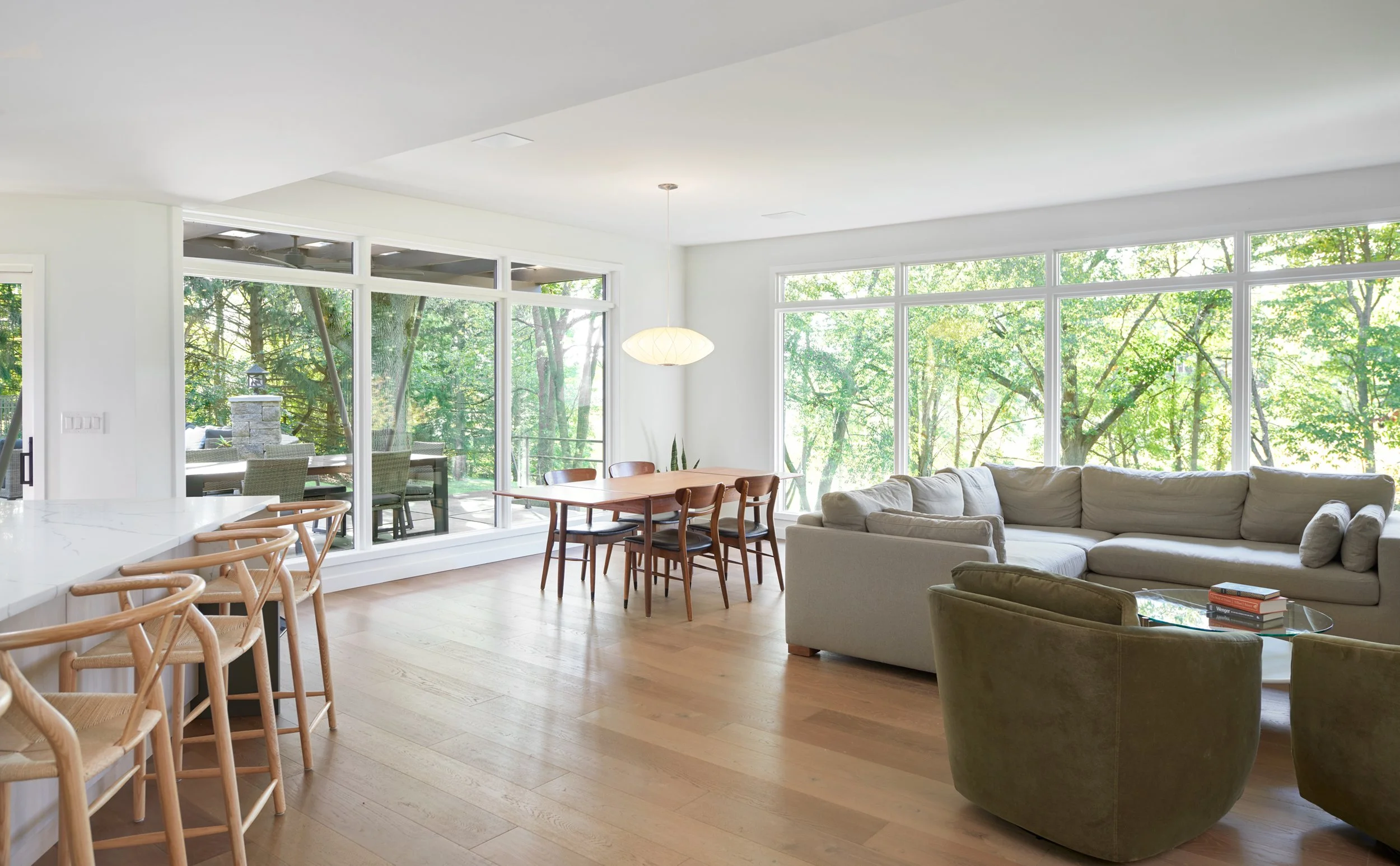 Open concept living area with large windows, light wood flooring, and wood-paneled ceiling contains beige sofa, and midcentury dining table in the background.