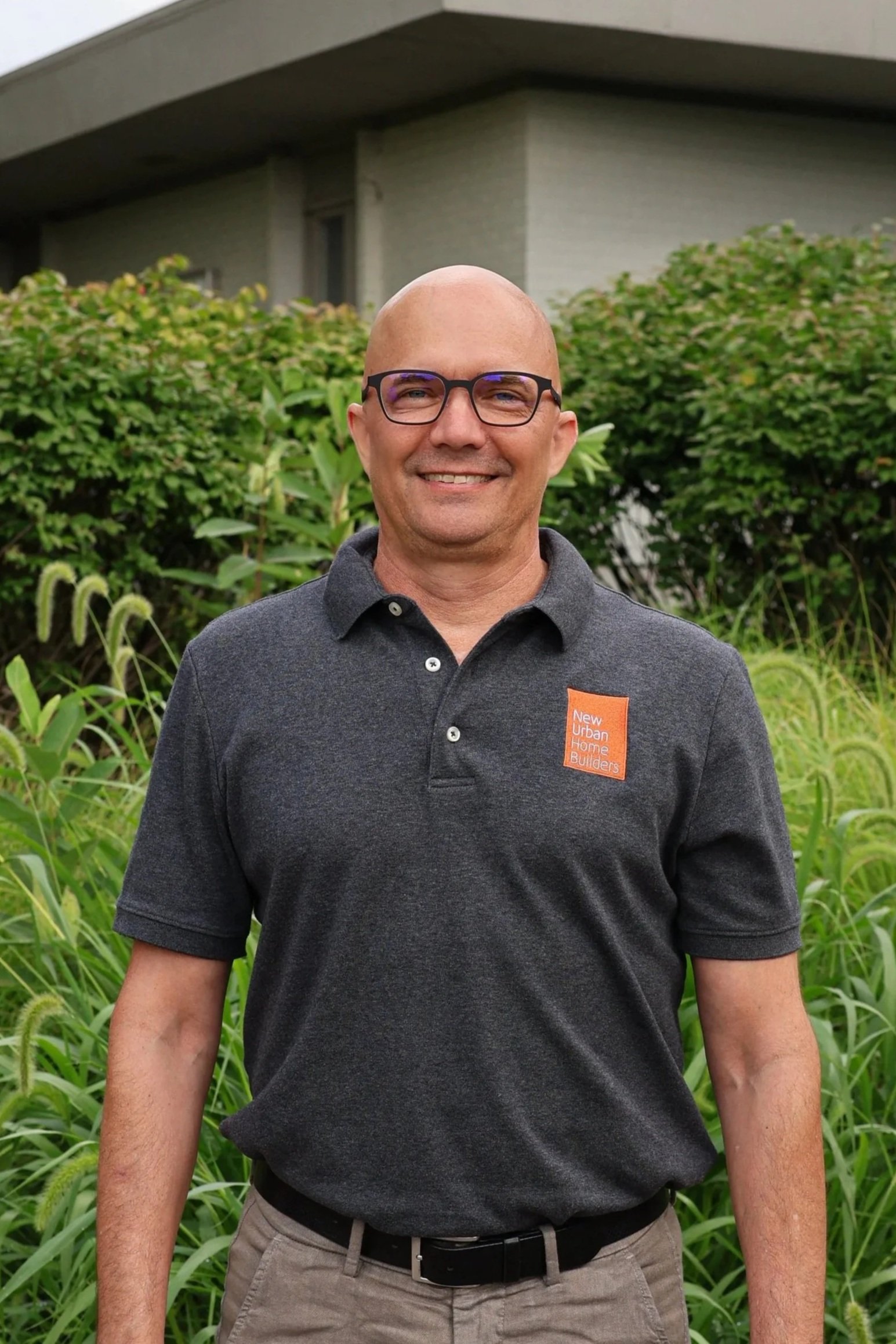Scott Branc in front of gray office building, wearing a dark gray polo shirt with an orange patch that says 'New Urban Home Builders'.
