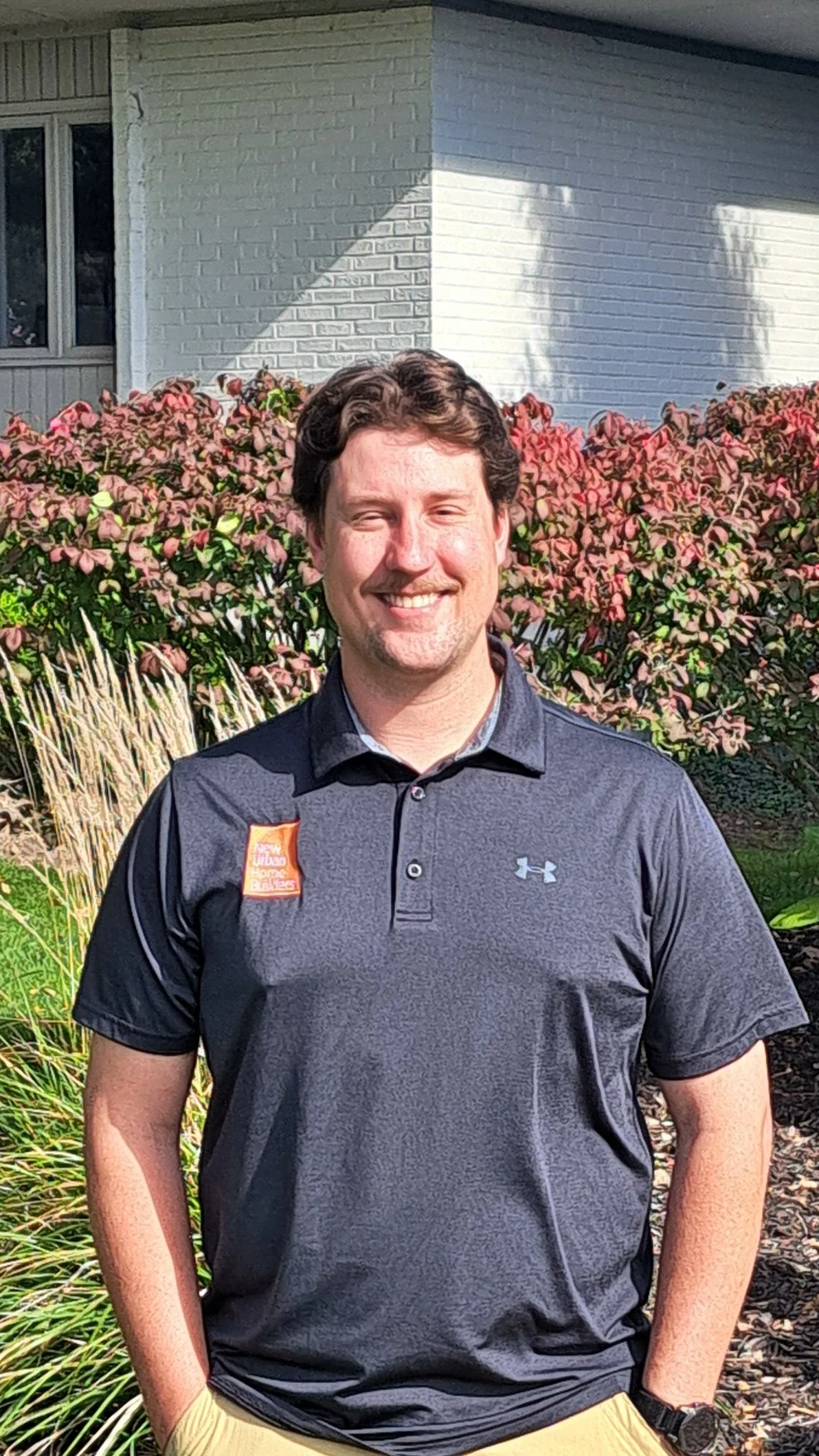 Blake Posthumus in front of gray office building, wearing a dark gray polo shirt with an orange patch that reads 'New Urban Home Builders'.