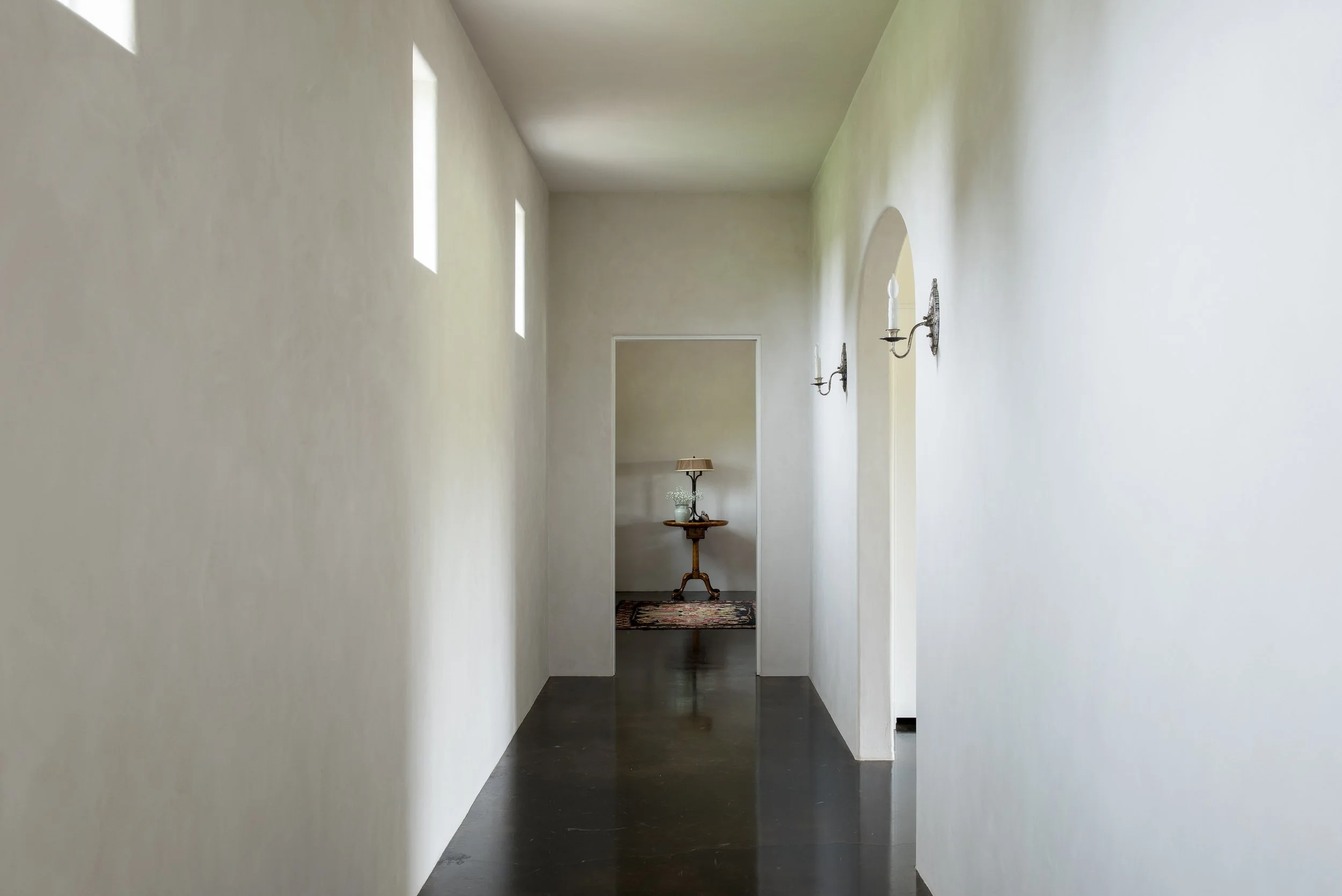 A minimalist hallway with white walls, small high windows on the left, black polished floor, wall-mounted candle-like sconces, leading to a room with a small table, a lamp, and decorative items.