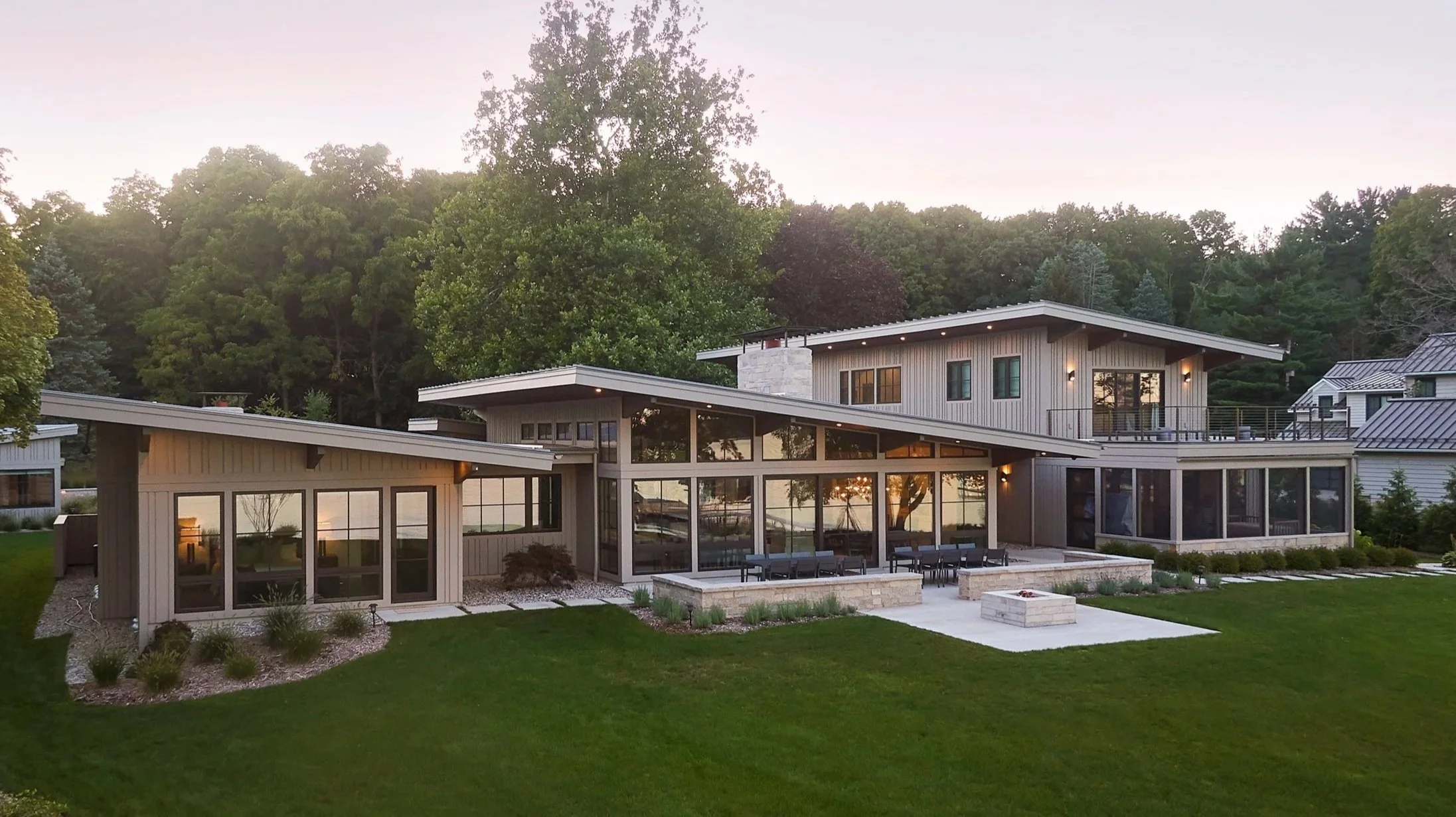 Modern two-story lakehouse with angled shed metal roofs, large glass windows, surrounded by green lawn and trees at dusk.