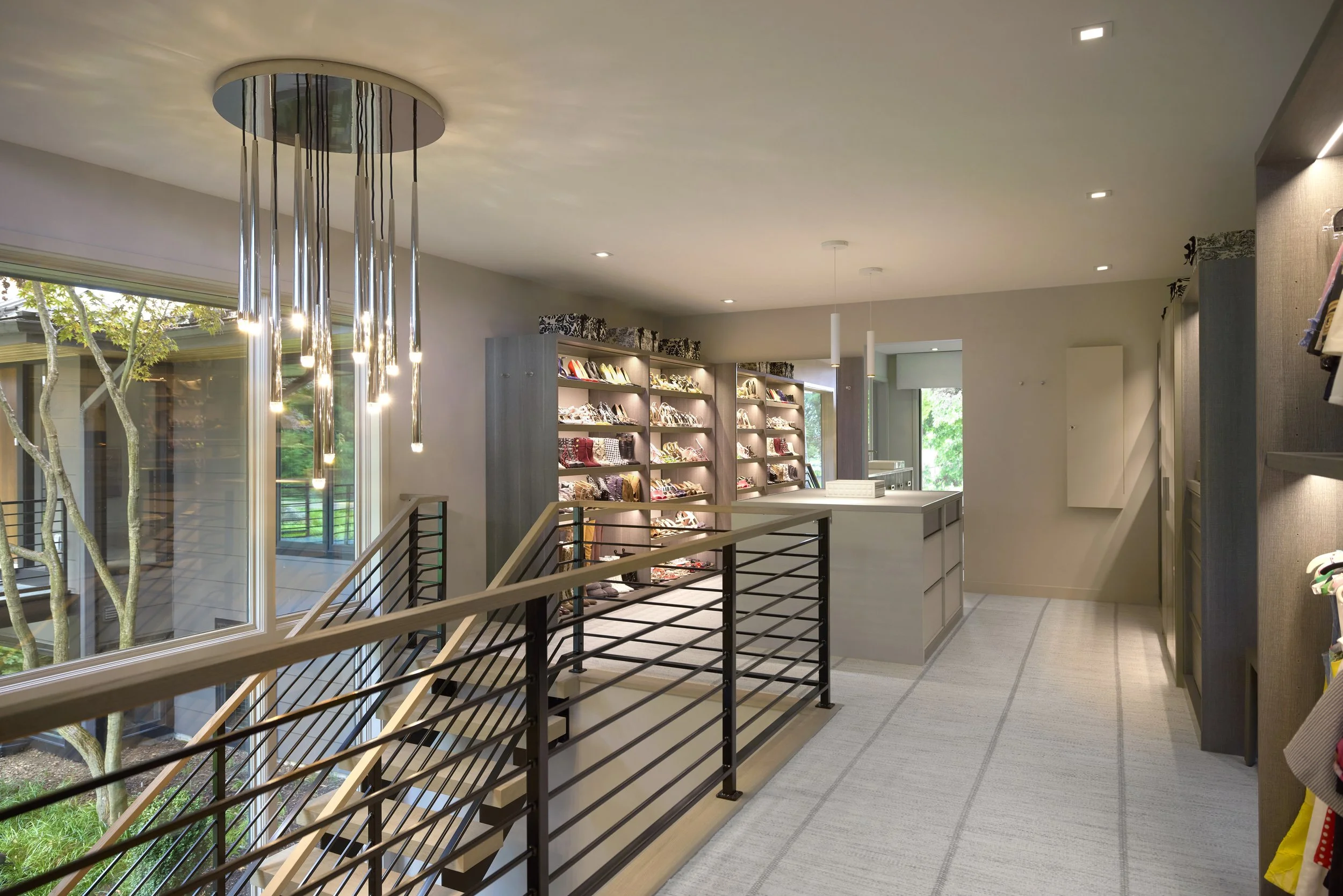Interior view of luxury master closet with handbags and accessories, illuminated modern lighting, large window showing outdoor trees, gray shelving, neutral-colored walls, and carpeted flooring.
