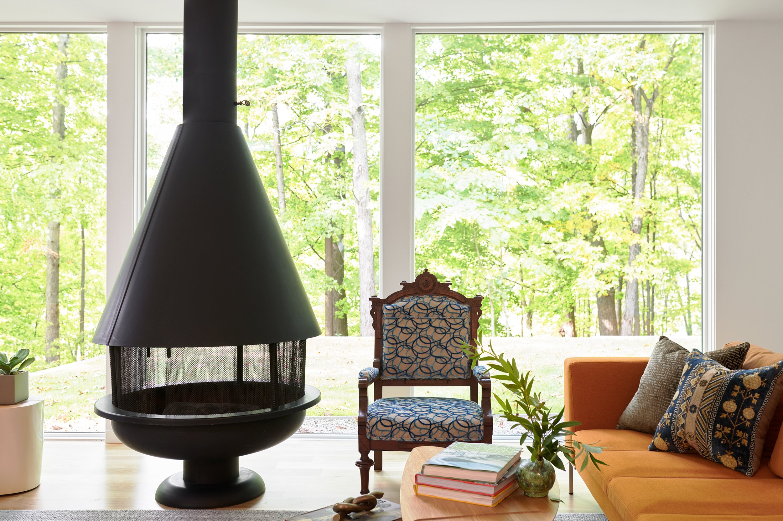 Living room with large window overlooking a green forest, a modern black fireplace, and furniture including a patterned armchair, orange sofa with decorative pillows, a small table with books, and a plant.