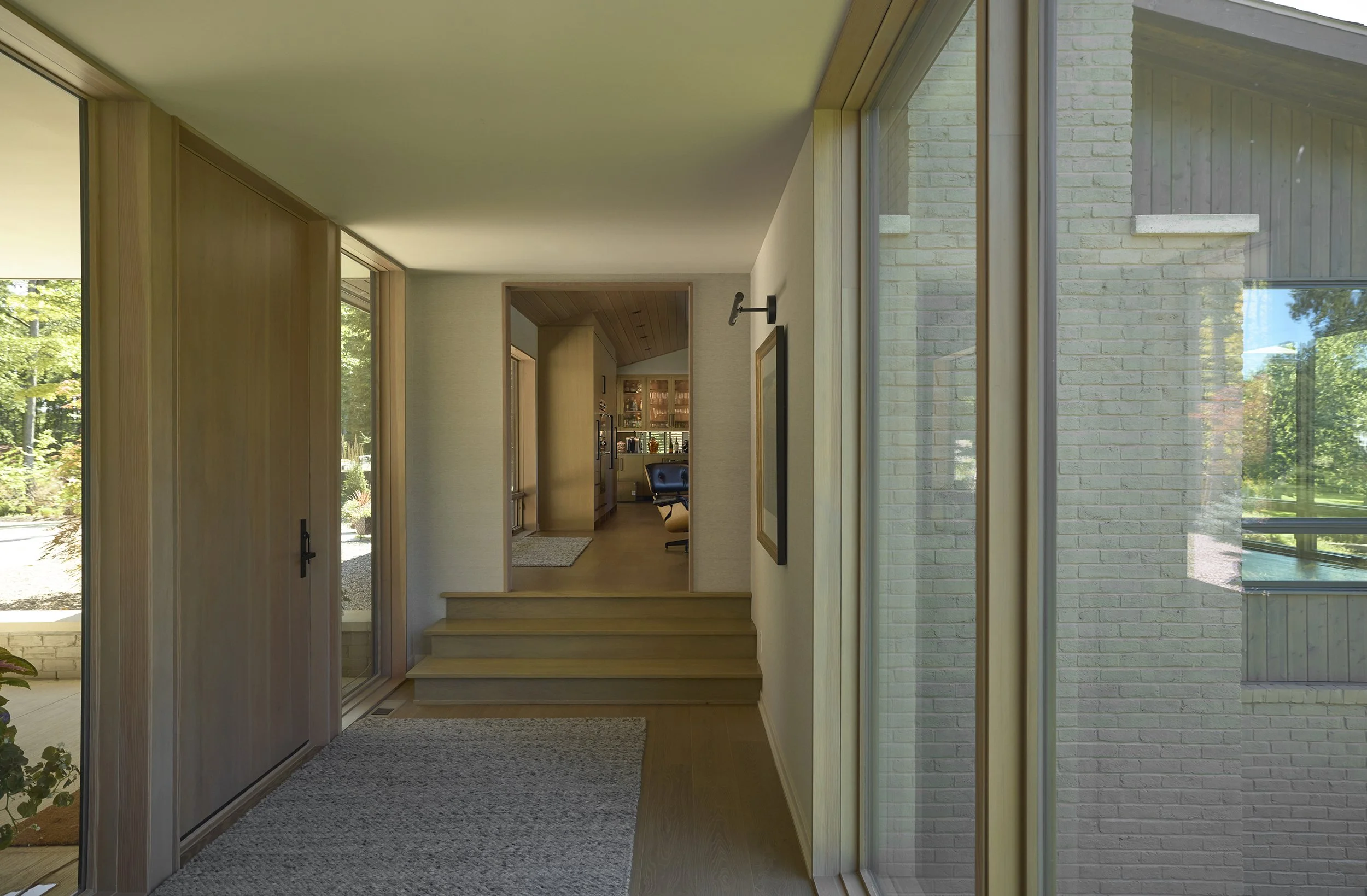 Interior hallway of a modern home with light wood flooring, a textured rug, steps leading to a room with a bar area containing shelves of bottles, and large windows allowing natural light inside.