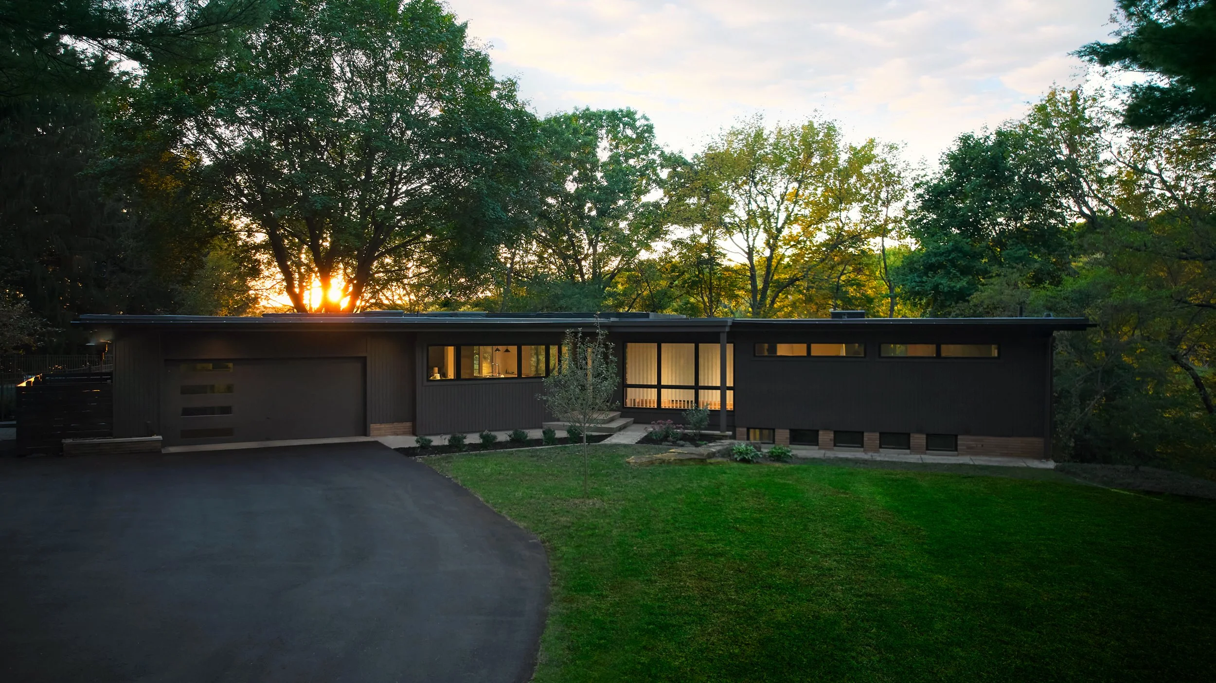 Midcentury home with flat roof and brown siding with a landscaped yard and setting sun in the background