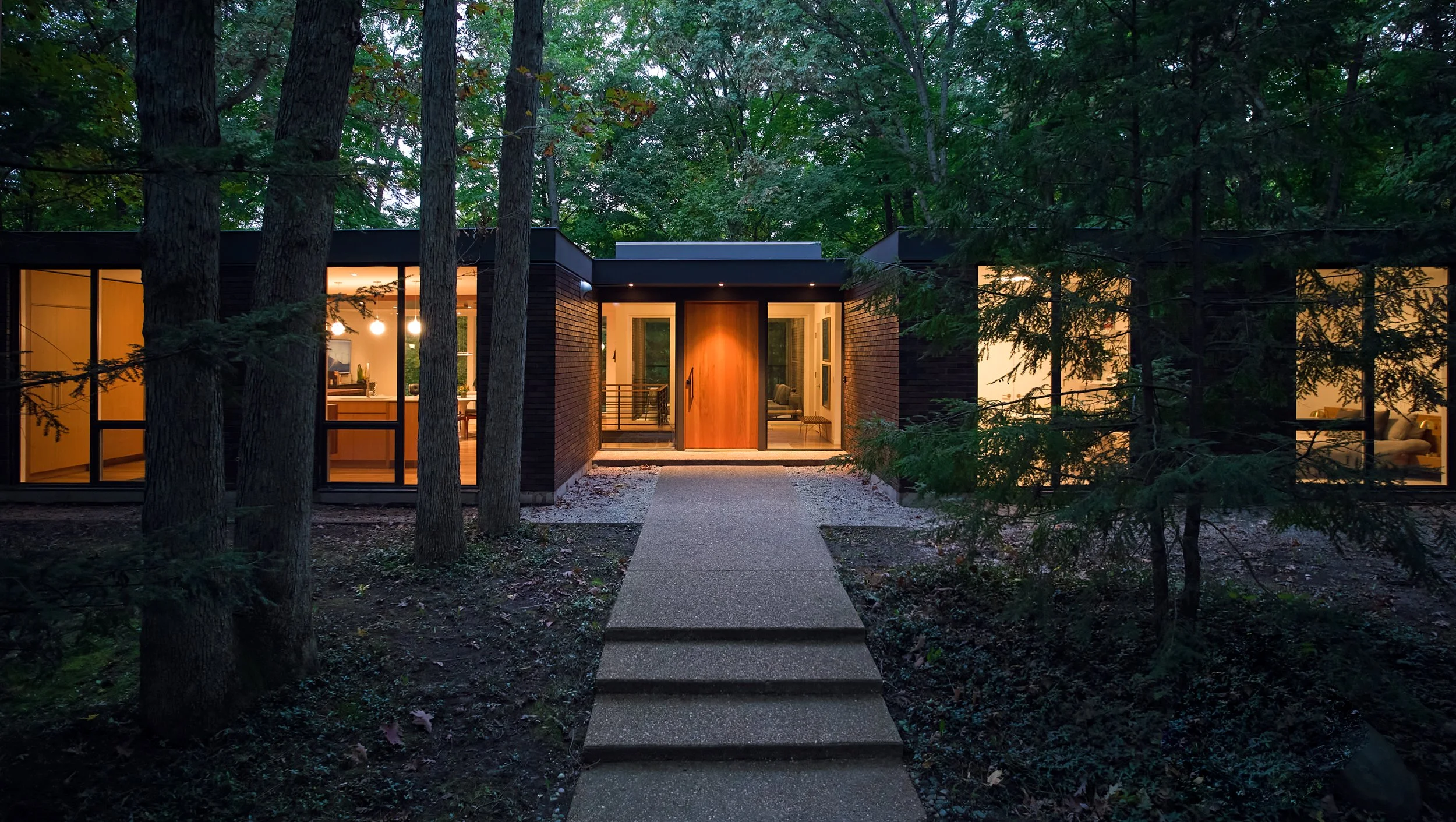 A midcentury modern house entryway with large glass windows illuminated from inside, situated in a wooded area at dusk, with a pathway leading to the front door.