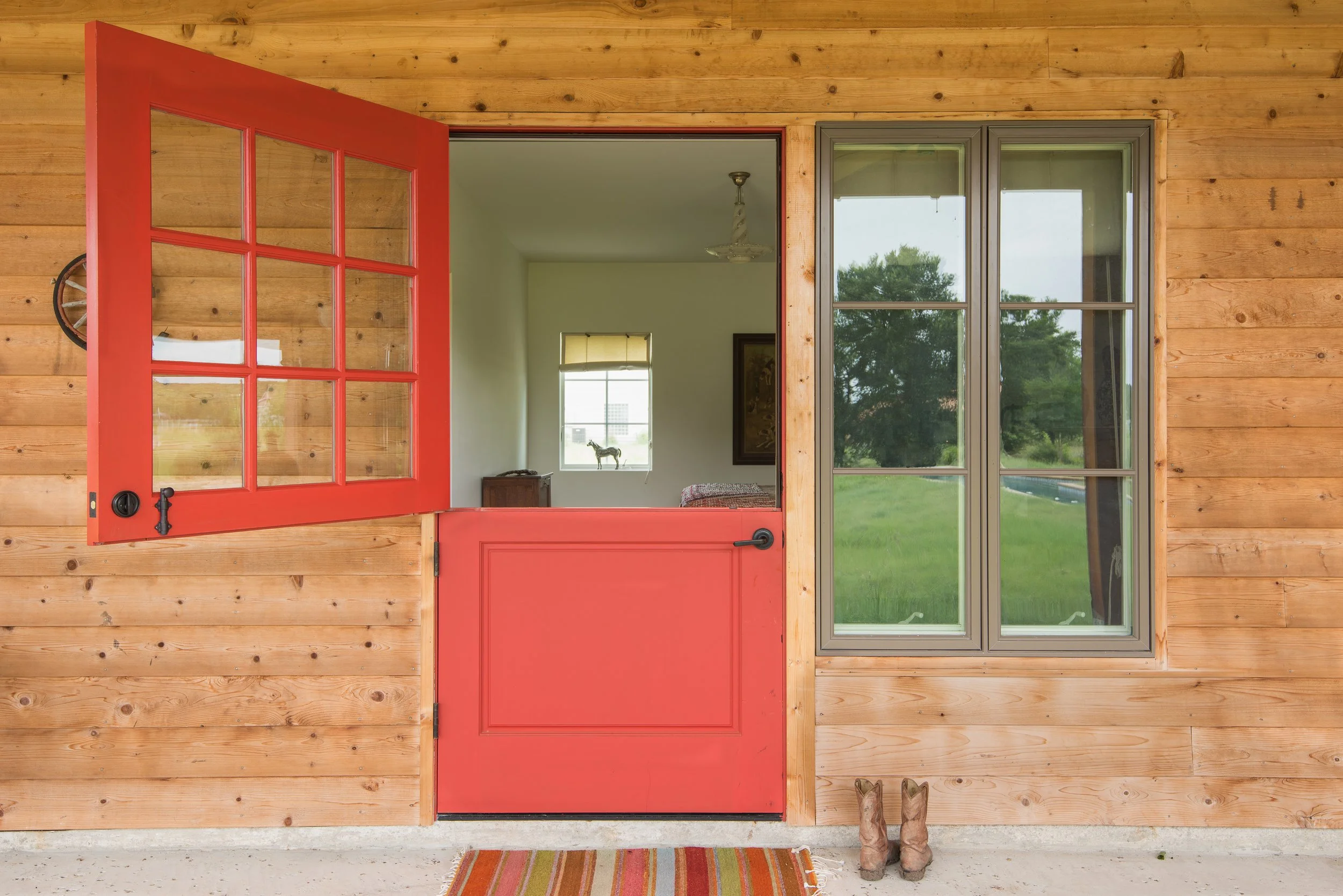A view through a red half door and window into a room with a small window, picture frame on the wall, and a horse figurine on a window sill. Outside the door, cowboy boots are on the ground and a striped rug is in front of the door.