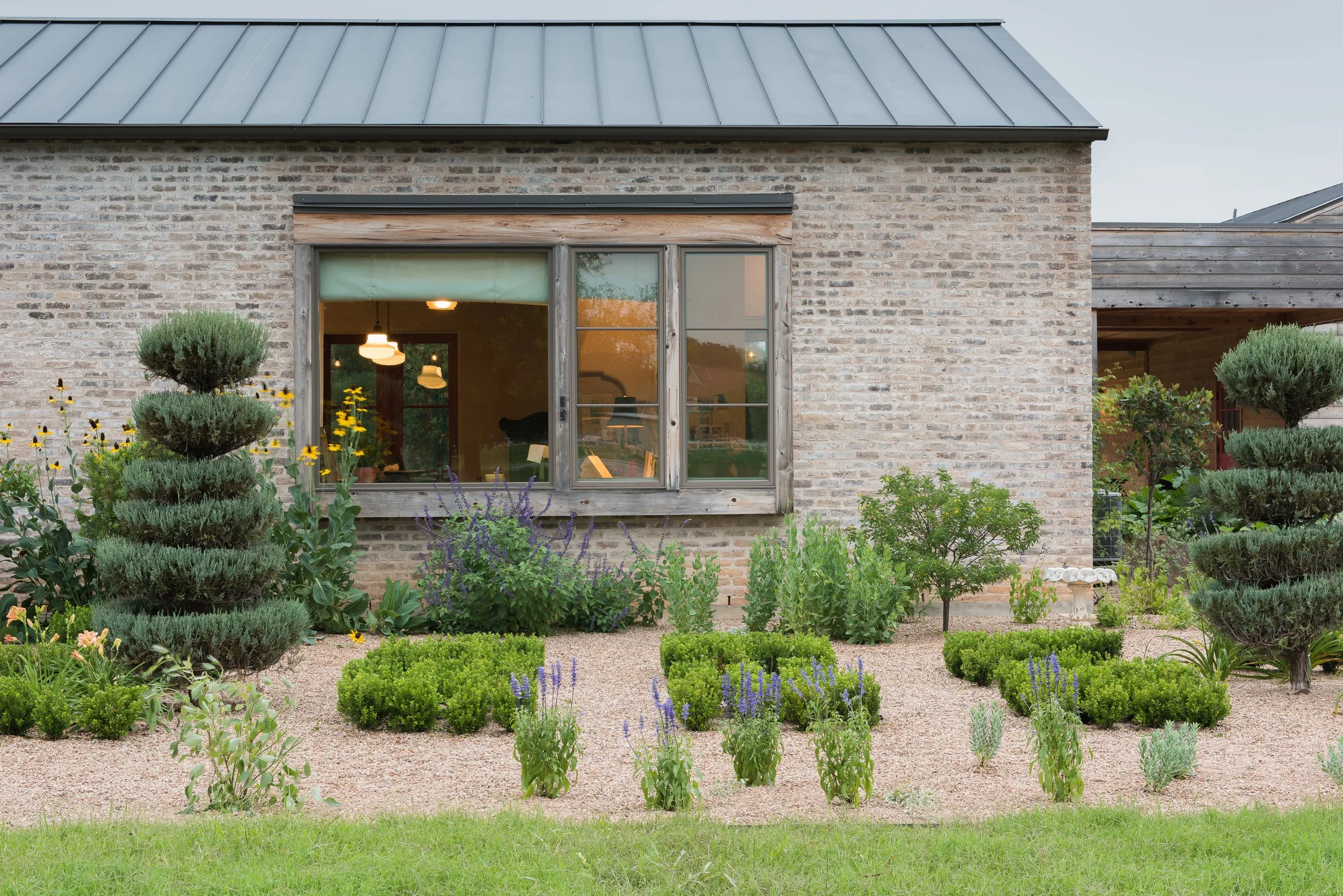 View of unique brick house with a large window, surrounded by a well-maintained garden with various shrubs, flowering plants, and small trees, and a gravel pathway in the front.