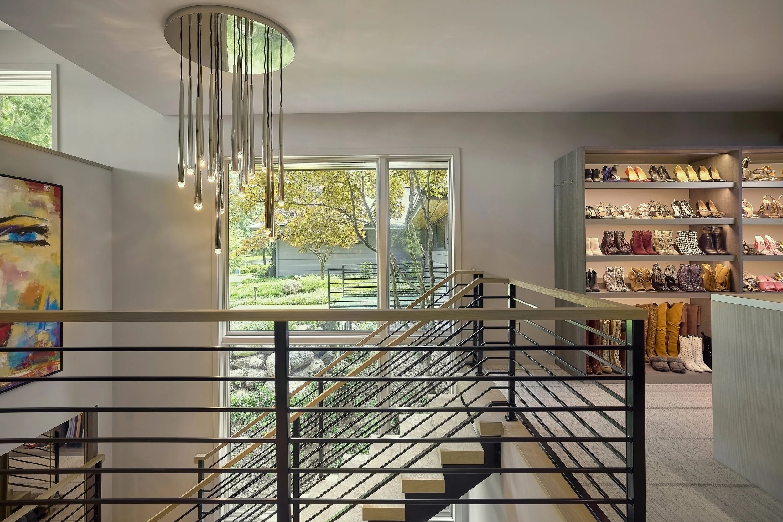 Interior view of high end master closet addition with a display of various footwear on shelves, large window showing trees outside, custom steel staircase and a decorative chandelier hanging from the ceiling.