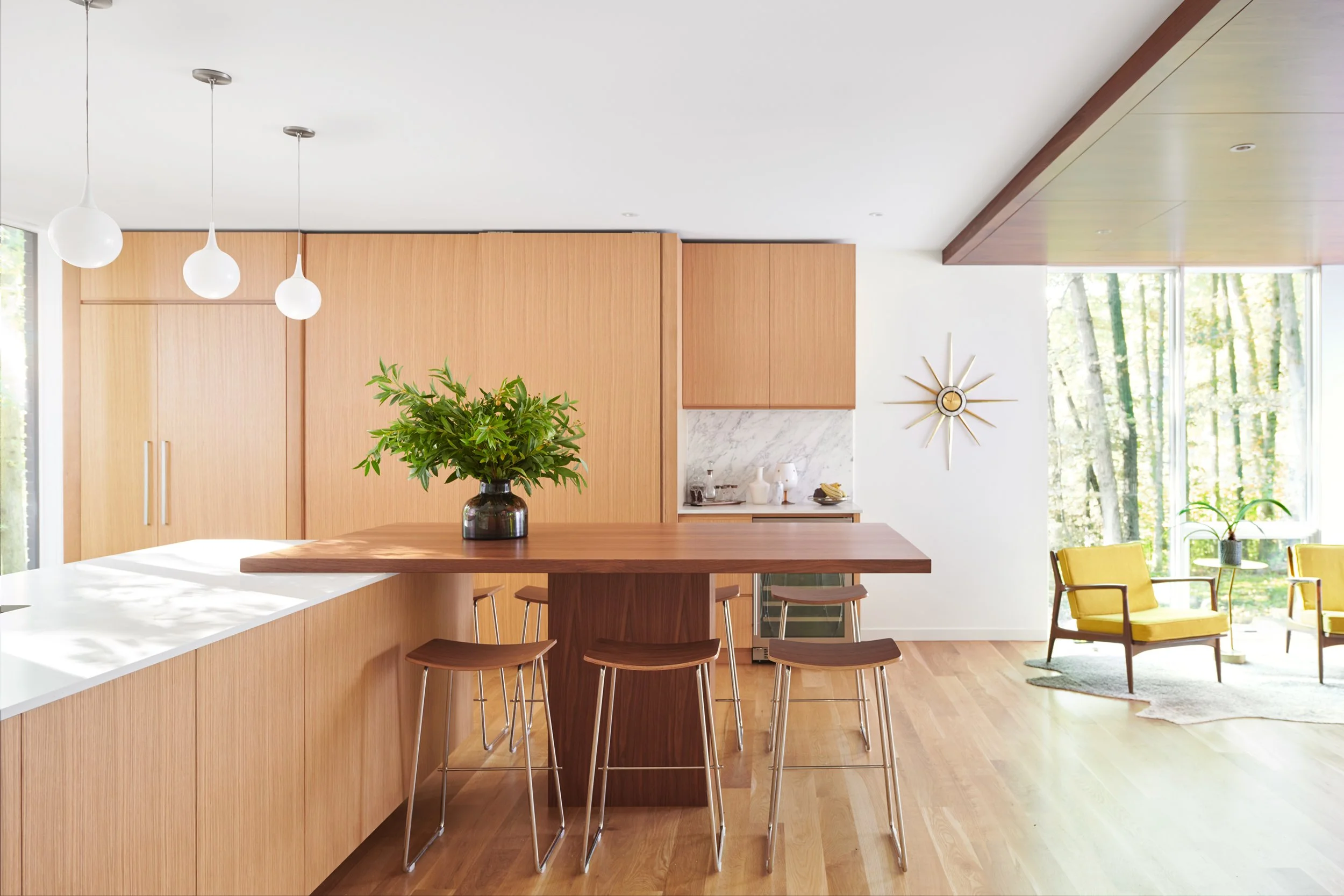 Modern kitchen with custom white oak cabinets, a kitchen island with bar stools, a large potted plant, and a sitting area with yellow chairs near large windows showing trees outside.