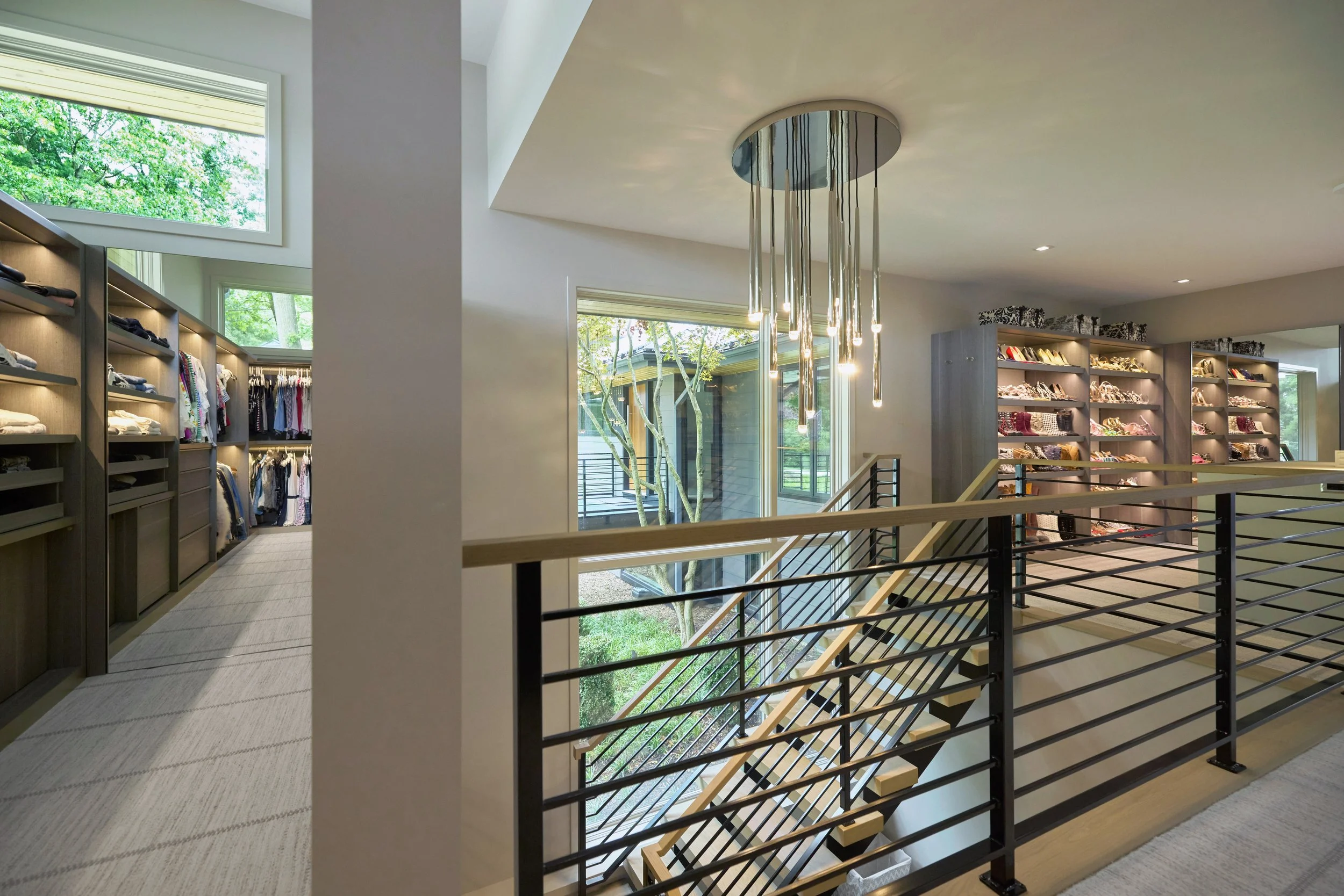 Modern interior of high end master closet with wool carpeted floors, statement ceiling light fixture, large windows showing green trees outside, and shelves filled with folded clothes and accessories.