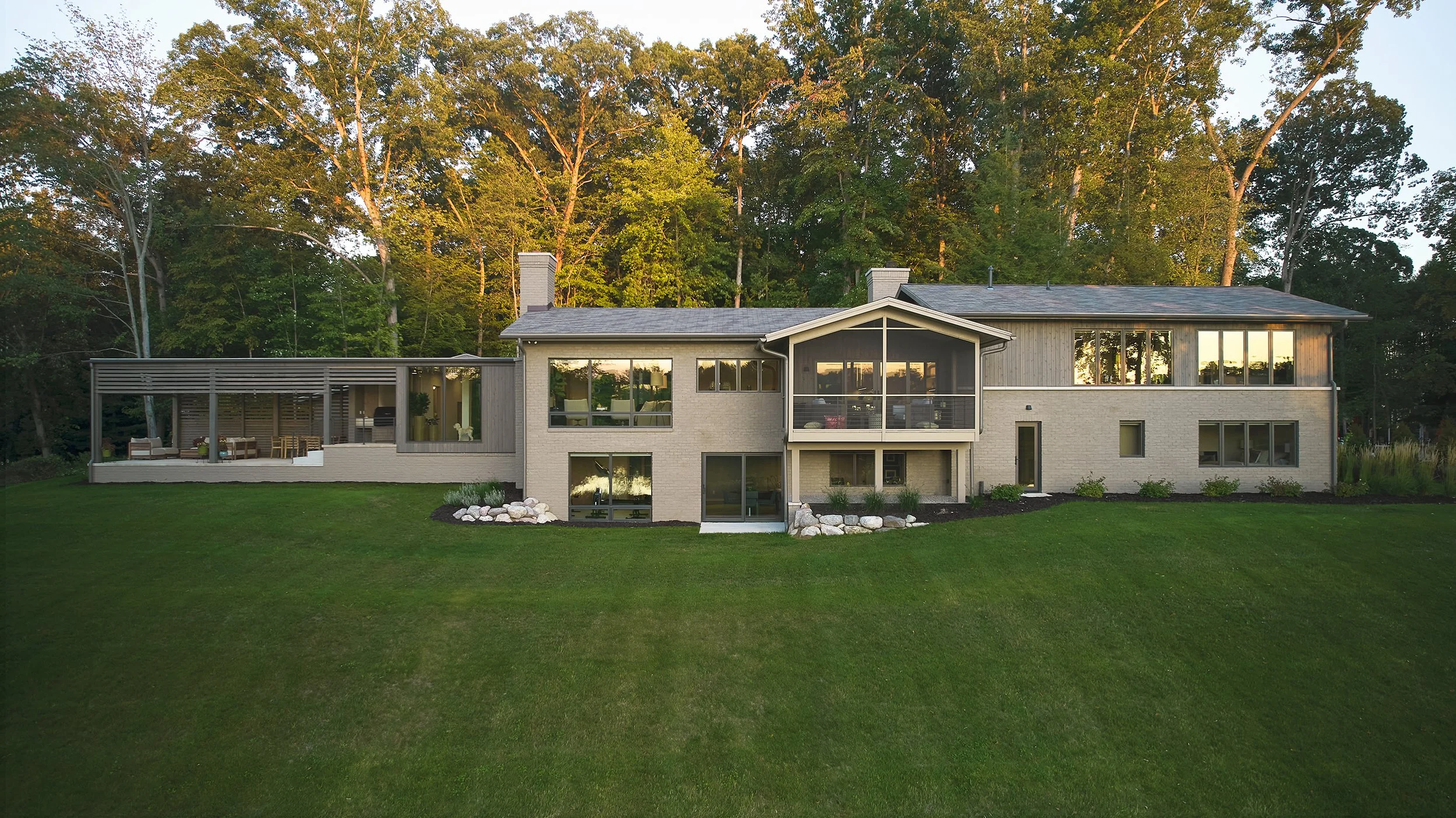 Rear view of waterfront modern house with walkout, screened porch, and well-maintained lawn, surrounded by trees.
