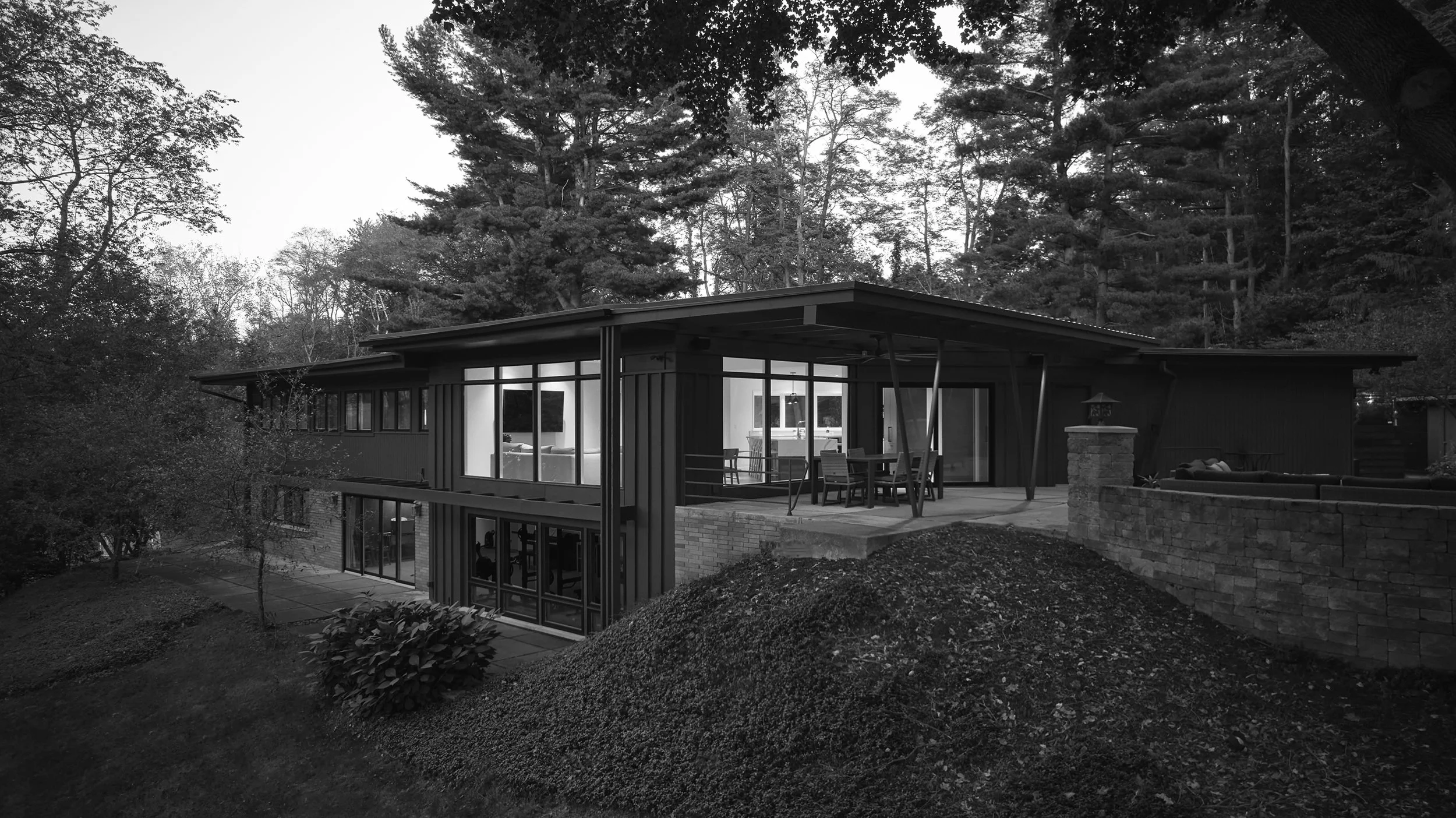 Black-and-white photo of a midcentury home's rear exterior.  An outdoor patio with dining table is featured.