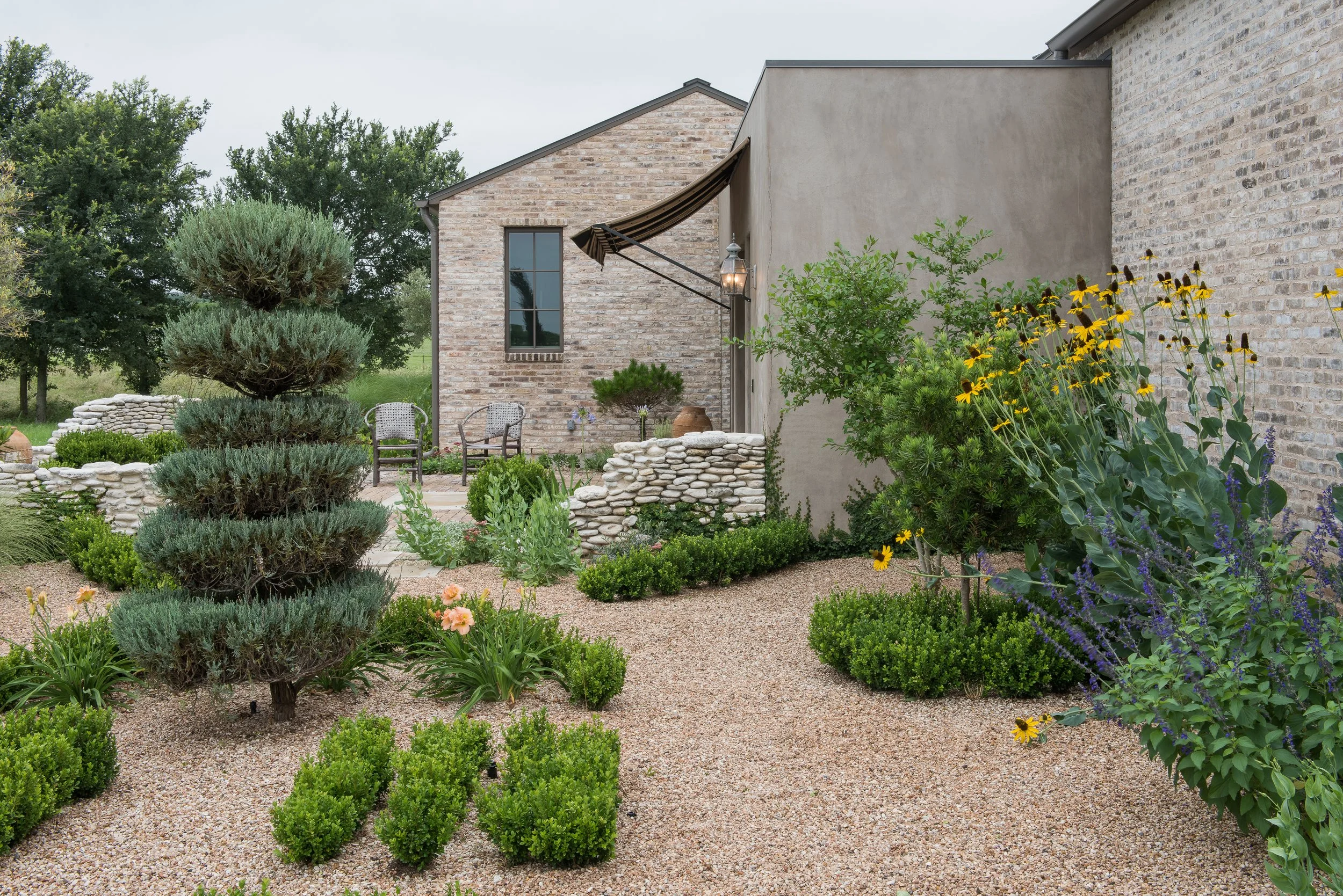 A landscaped backyard garden with a trimmed tree, bushes, yellow flowers, stone borders, chairs, and a house with brick walls and a window.