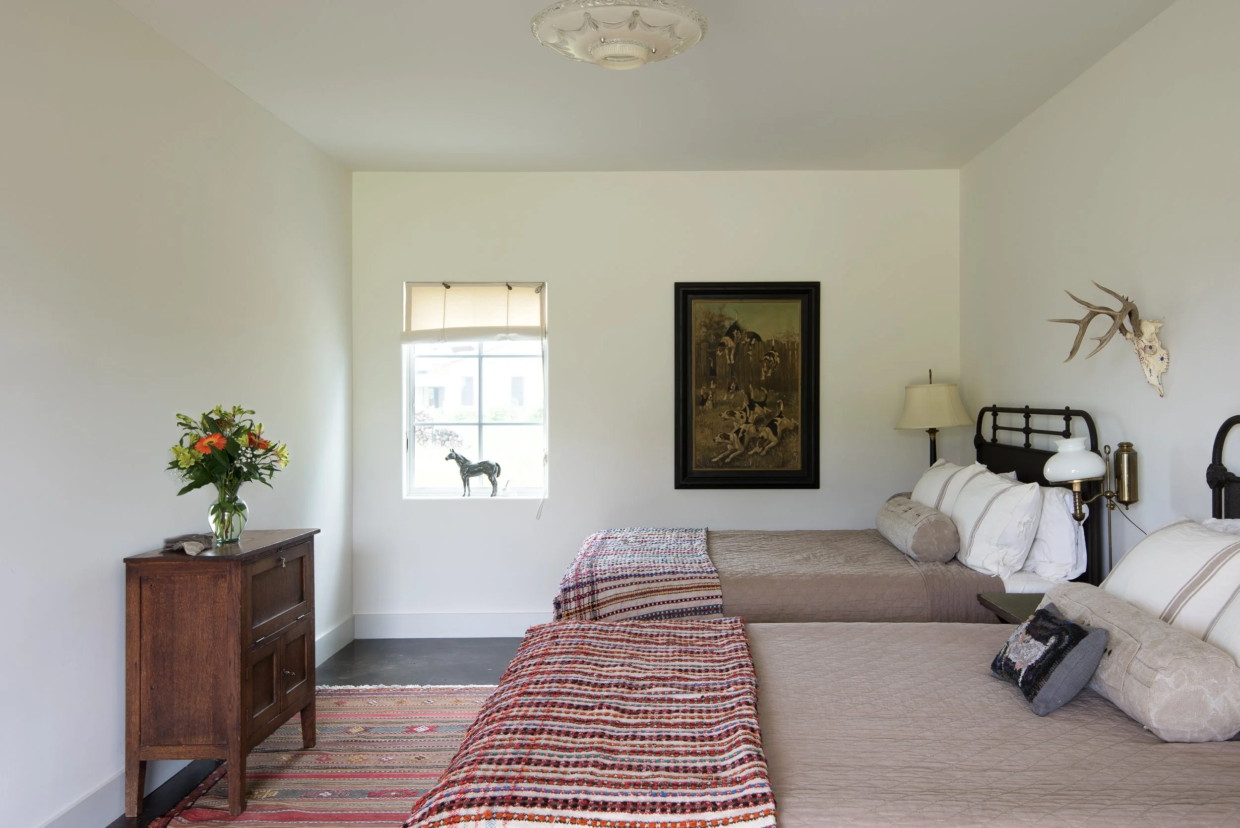 Guest bedroom with two beds, a window with a small horse figurine, a picture on the wall, a lamp, deer antler wall decor, a vase of flowers, a small piece of art on the bed, and a colorful striped rug.