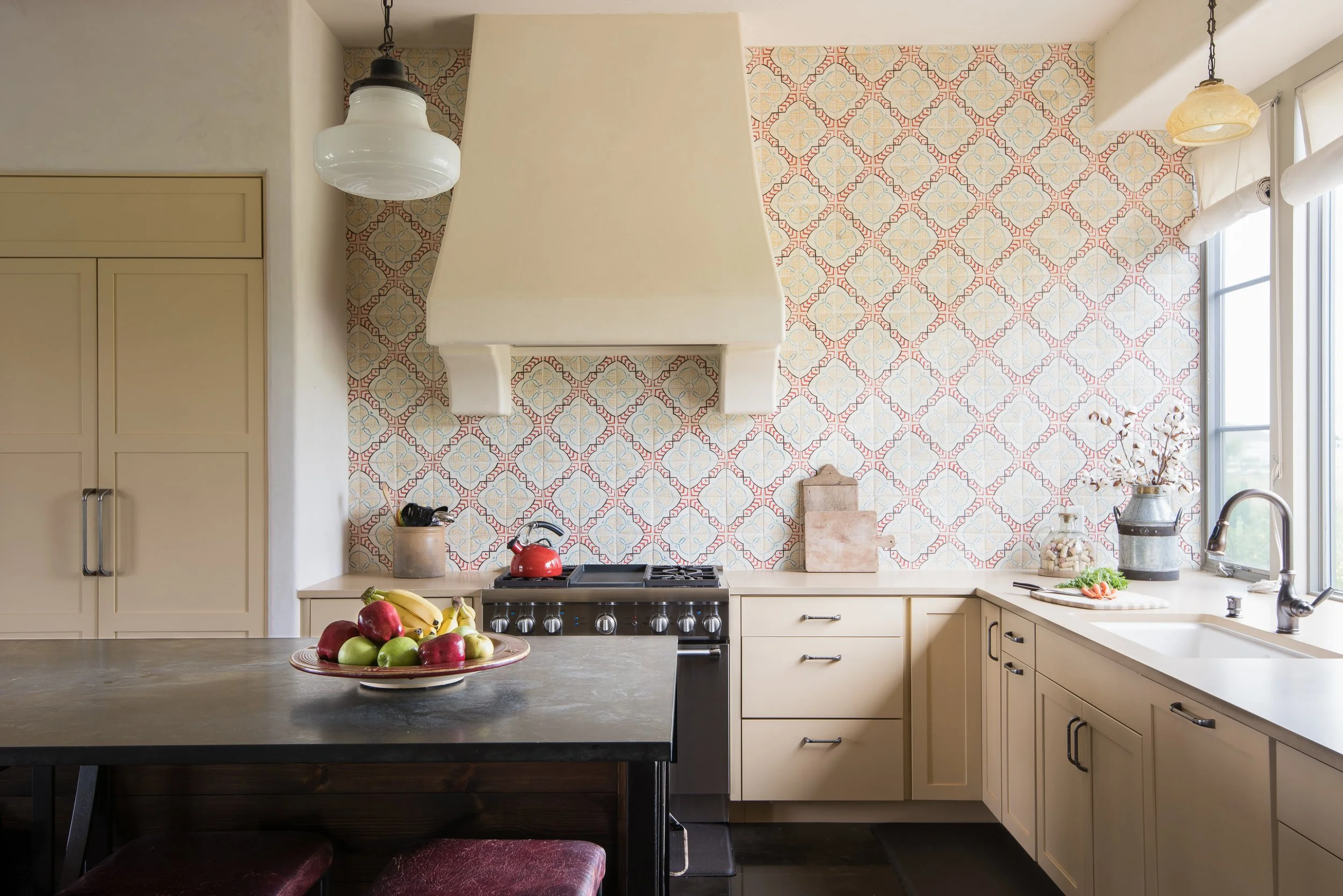 A kitchen with beige cabinets, a black countertop, a stove with a red kettle, a fruit bowl with apples and bananas, a beige range hood, a tiled backsplash with a colorful pattern, a white window with a view outside, a farmhouse sink, and various decorative items.