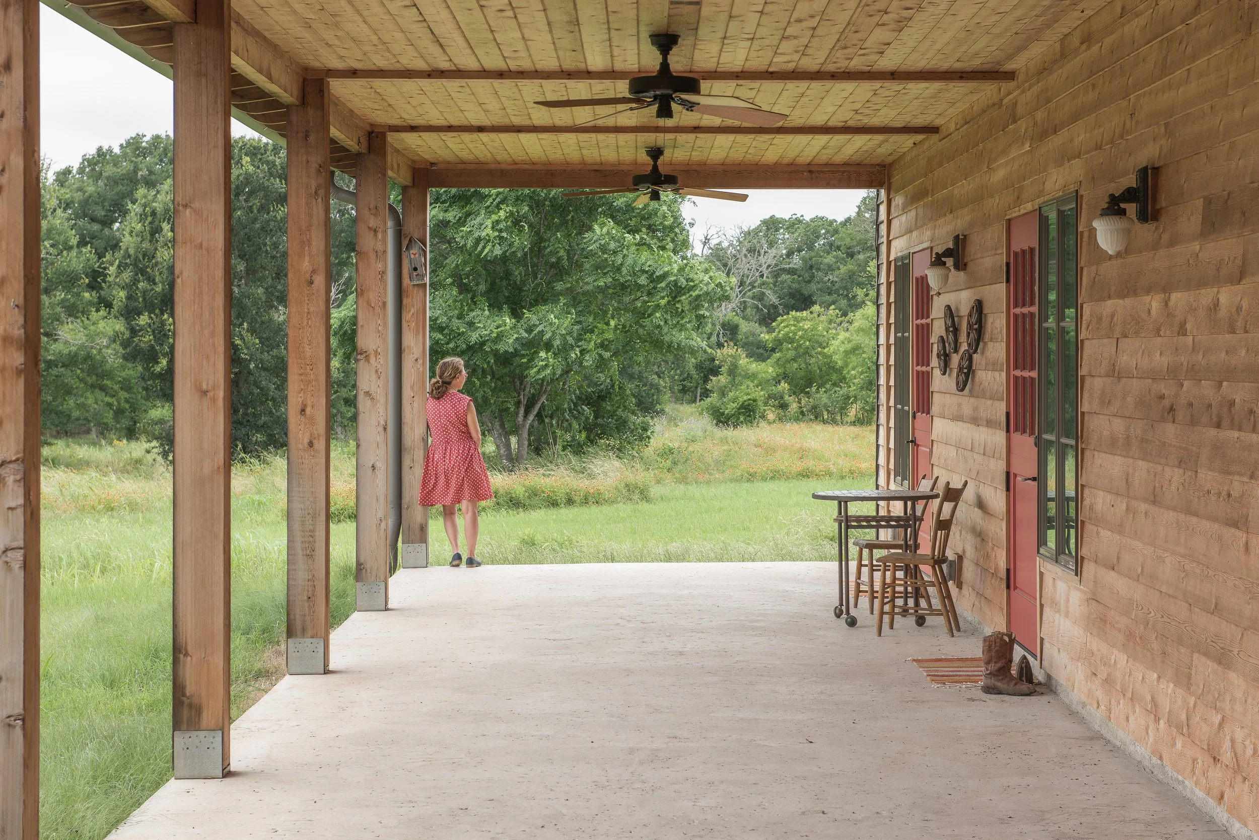 A woman in a pink dress standing on a porch looking out at green field and trees.