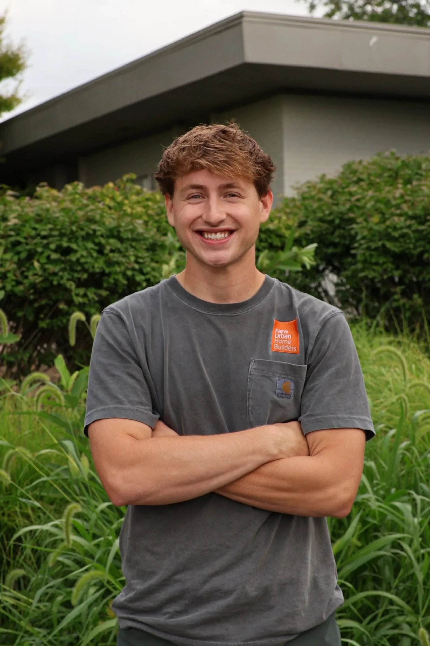 Jake Wilgus in front of gray office building, wearing a gray T-shirt with a pocket that has an orange patch reading 'New Urban Home Builders'.