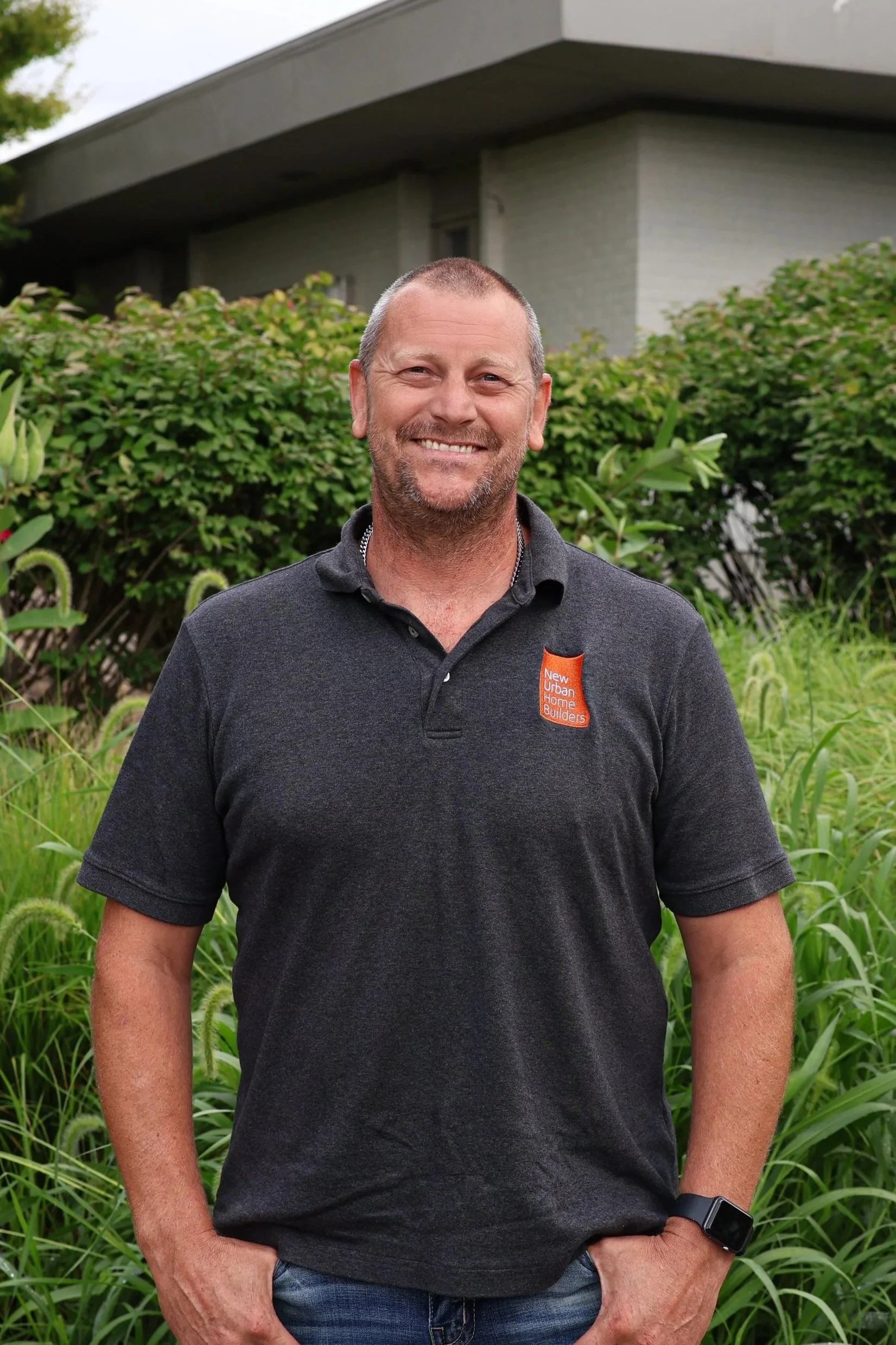 Brandon VanNoord in front of gray office building, wearing a dark grey polo shirt with an orange patch that says 'New Urban Home Builders'.